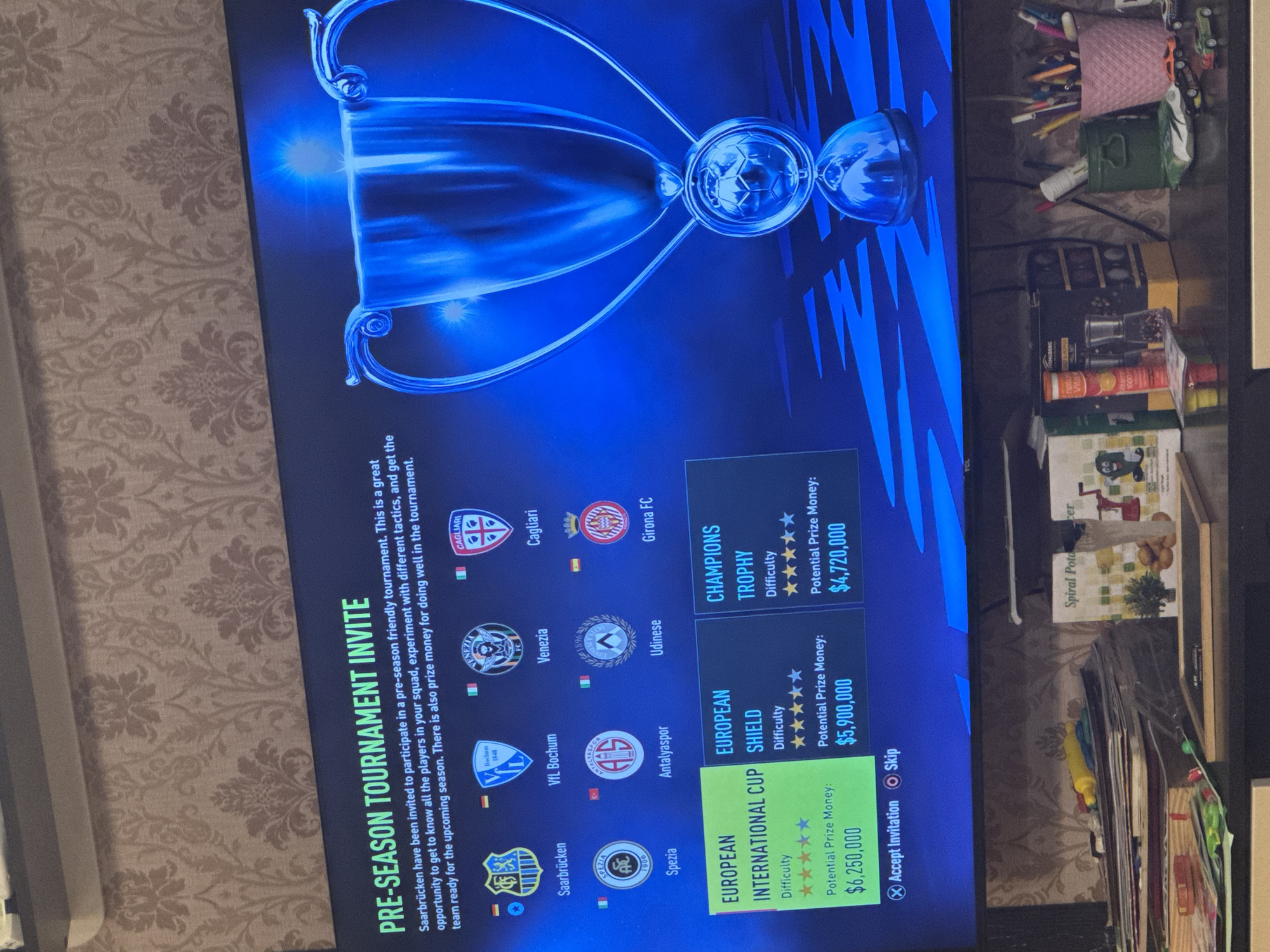 The image shows a television screen displaying a video game menu. The game is FIFA and the menu is for pre-season tournament invites. Three tournament options are presented: European International Cup, European Shield, and Champions Trophy, each with a different prize money and difficulty rating. The player has highlighted the "European International Cup" with a potential prize money of $6,250,000 and a four-star difficulty rating.
The game displays logos of the teams participating in the pre-season tournament, including Saarbrücken, Spezia, VfL Bochum, Antalyaspor, Venezia, Udinese, Cagliari, and Girona FC.
In the background, outside the television screen, is a patterned wallpaper, possibly cream or light brown in color. To the right of the screen, there's a cluttered area with various items, including a pink cup holding pens, a green container, boxes of supplements, and a spiral potato slicer box.