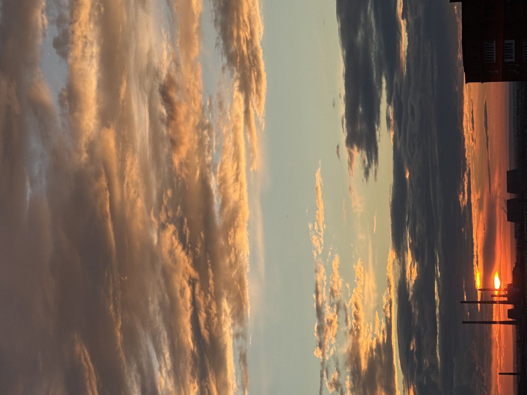 The image captures a sunset over an industrial cityscape. The sky is filled with dramatic clouds, painted in hues of orange, gold, and grey, with streaks of blue peeking through. In the foreground, silhouetted against the vibrant sunset, are several tall industrial smokestacks, suggesting a factory or power plant. To the right, the corner of a brick building is visible, hinting at residential or commercial structures within the city. The overall impression is one of an industrial area experiencing the beauty of a sunset.