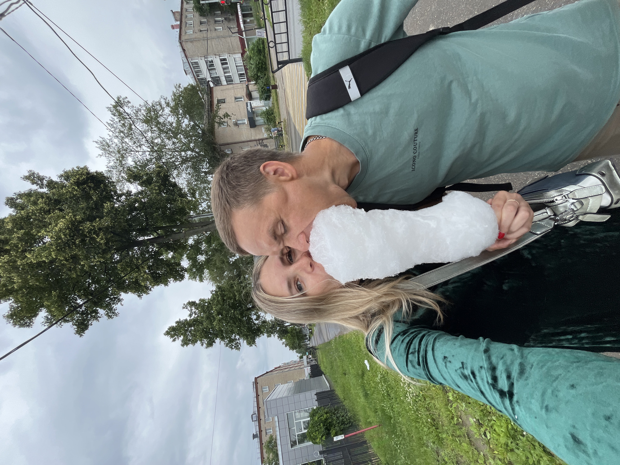 A selfie is taken outdoors on a cloudy day in the city of Chaikovsky, Russia. A woman with blonde hair is on the left, wearing a green velvet dress. A man is on the right, wearing a teal t-shirt with a black backpack. They are both holding a large piece of white cotton candy, with the man taking a bite out of it. The background shows a sidewalk, grass, trees, and apartment buildings, consistent with an urban environment in Russia.