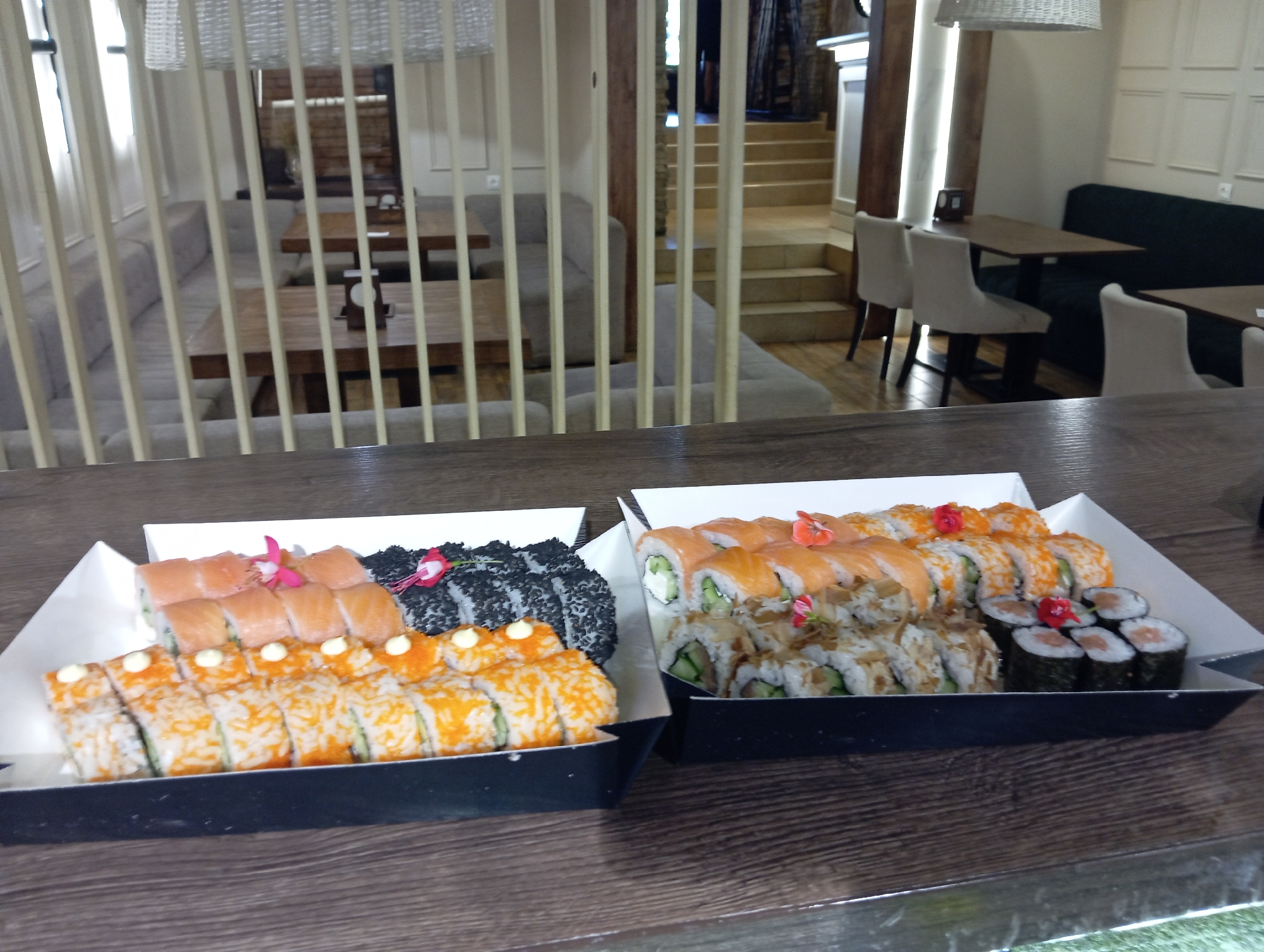 The image shows two open sushi boxes placed on a wooden table. The sushi boxes contain various types of sushi rolls. One box on the left has orange roe-covered rolls and rolls topped with white sauce. The box on the right has salmon-topped rolls, rolls covered in black sesame seeds, and rolls with cucumber and cream cheese. The background suggests an indoor setting, possibly a restaurant or cafe, with tables, chairs, and decorative elements such as wooden beams and lighting fixtures. The overall scene is a close-up view of the sushi presentation, with the restaurant interior blurred in the background. There are no people visible in the image.