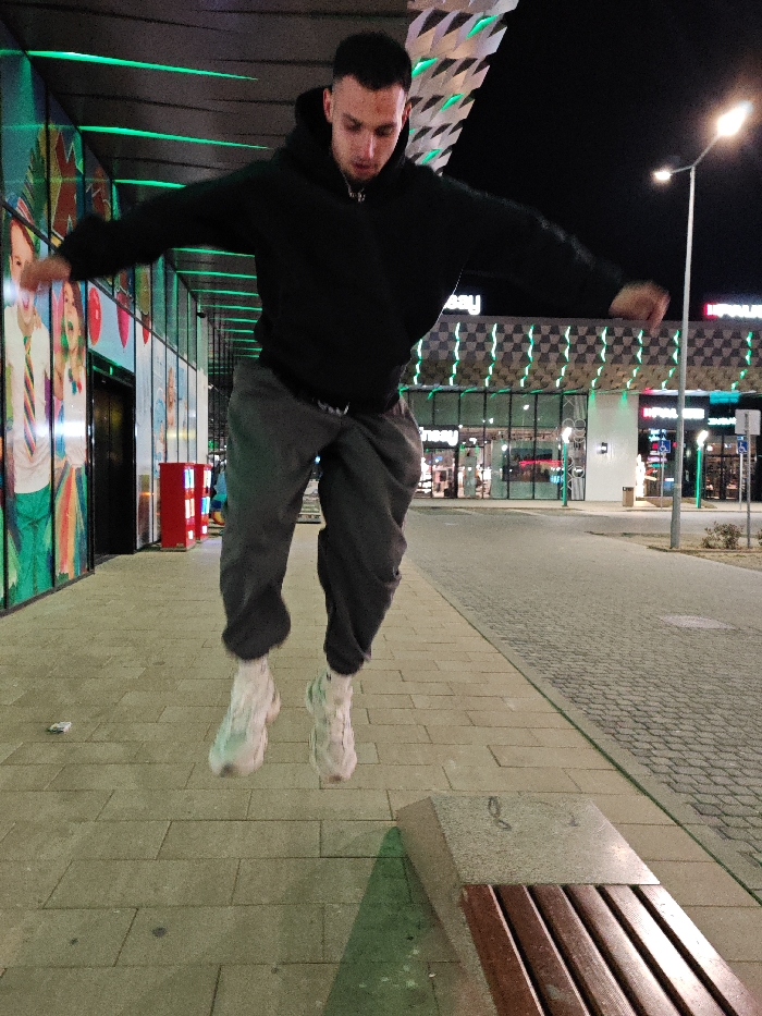 The image depicts a young adult male in mid-air during a jump on a paved pedestrian walkway at night, outside the Grand Mall Shumen in Shumen, Bulgaria.
The male subject, appearing to be in his 20s with short dark hair, is wearing a black hooded sweatshirt, dark grey sweatpants, and white athletic shoes with chunky soles. His arms are outstretched horizontally, and his body is slightly forward-bent, indicating an active leap. His facial expression appears focused downwards.
The immediate foreground features light-colored rectangular paving stones. A wooden slatted public bench with a concrete end support is visible in the lower right, partially cropped. A small piece of white debris rests on the pavement near the subject's left foot.
The background is dominated by the illuminated facade of the Grand Mall Shumen. This modern commercial building features large glass panels, some displaying colorful graphic advertisements. A distinctive architectural element of the building is a pattern of horizontal green LED lights, arranged in a checkered fashion across its upper sections. Visible signs for "Kaufland" and "Intersport" confirm the commercial nature of the building. A tall streetlamp with a bright light source stands on the right, and a red vertical object, possibly a utility box or vending machine, is against the building's glass facade on the left. The area beyond the walkway appears to be a cobbled surface, likely a road or parking lot, under artificial light.