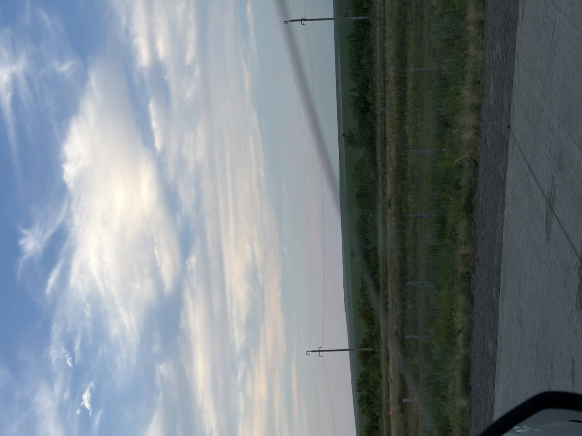 The image captures a view of a rural landscape under a cloudy sky. In the foreground, a paved surface, possibly a road or pathway, is visible. Beyond this, a strip of dry grass and a fence with wooden posts delineate a field. Further in the background, there are rows of green vegetation, likely crops or trees, with utility poles and overhead wires stretching across the scene. A dark, smoky plume extends diagonally across the sky from the right side of the image towards the center-left, obscuring a portion of the distant landscape. The overall impression is one of a vast, open area with signs of human infrastructure and a natural phenomenon in the sky. The specific location within Баймырза, Kazakhstan, is not discernible from the visual elements present.