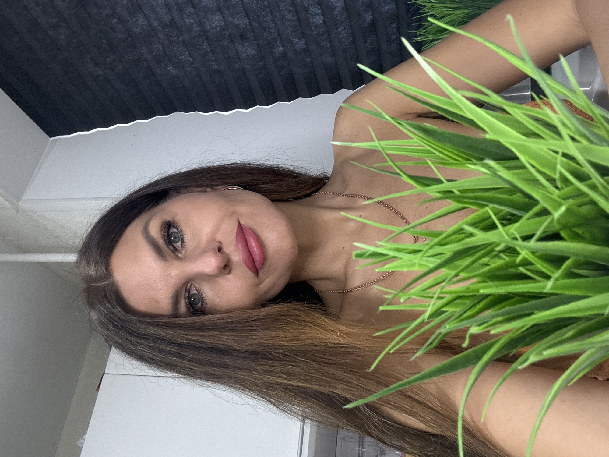 A woman is posing indoors with green, artificial grass obscuring the right side of the image. She is fair-skinned with long brown hair, visible jewelry, and makeup. The background features a white wall and a dark gray roller blind. The location is presumed to be an indoor space, possibly in the city of Chaikovsky, Russia.