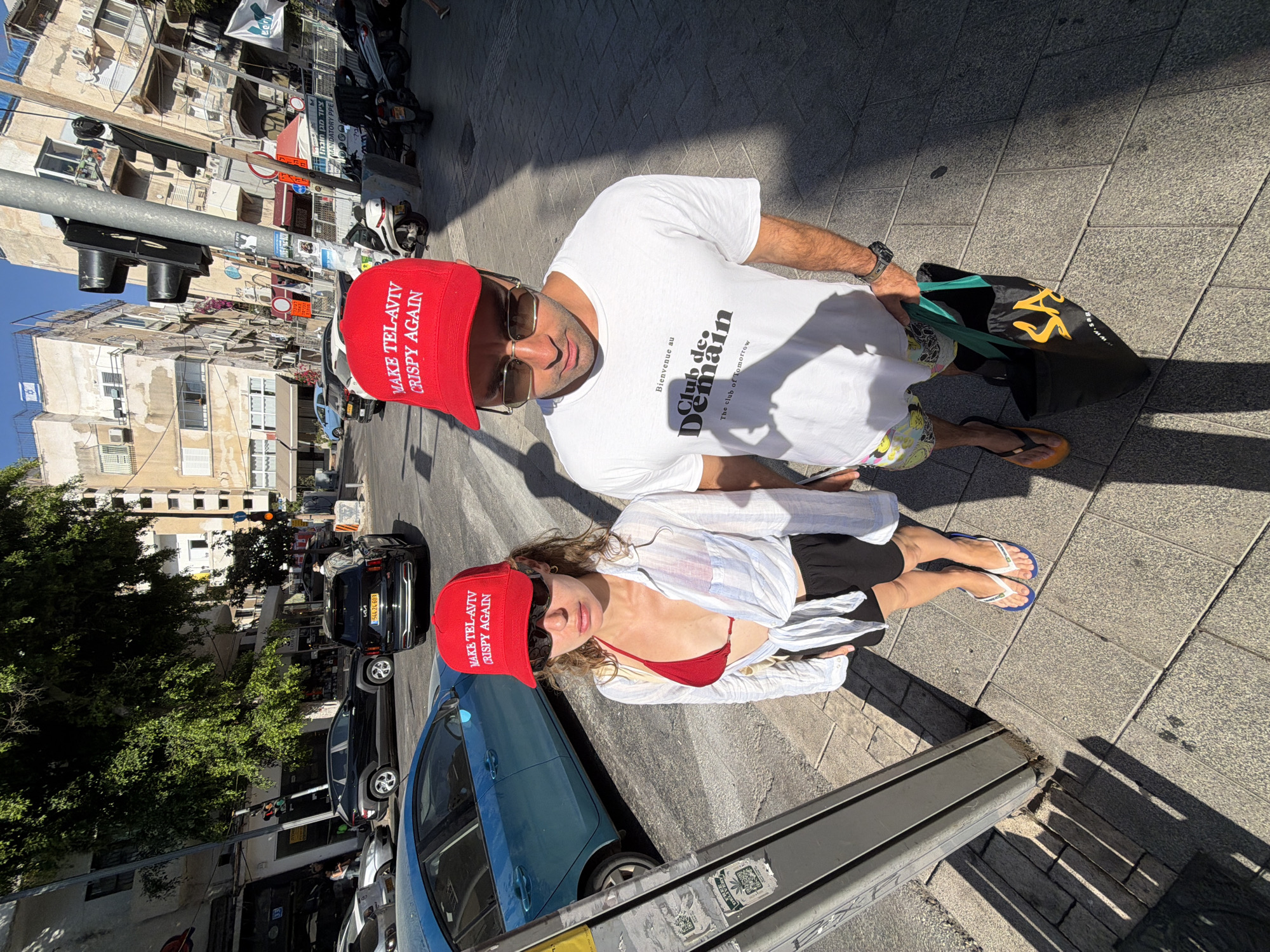 A man and a woman stand on a sidewalk in Tel Aviv, Israel, under a clear sky. The woman, on the left, wears a red bikini top, a white unbuttoned shirt, black shorts, and blue flip-flops. She has curly brown hair and is wearing sunglasses. On her head, she wears a red cap with white text that reads "MAKE TEL-AVIV CRISPY AGAIN". The man, on the right, wears a white t-shirt with black text that reads "Bienvenue au Club de Demain. The club of Tomorrow", a colorful patterned pair of shorts, sunglasses, and orange flip-flops. He holds a black tote bag with a green handle. A traffic light is visible to the left of the couple, and various buildings and parked vehicles, including a blue car and black SUVs, line the street. The sun casts long shadows on the paved sidewalk.