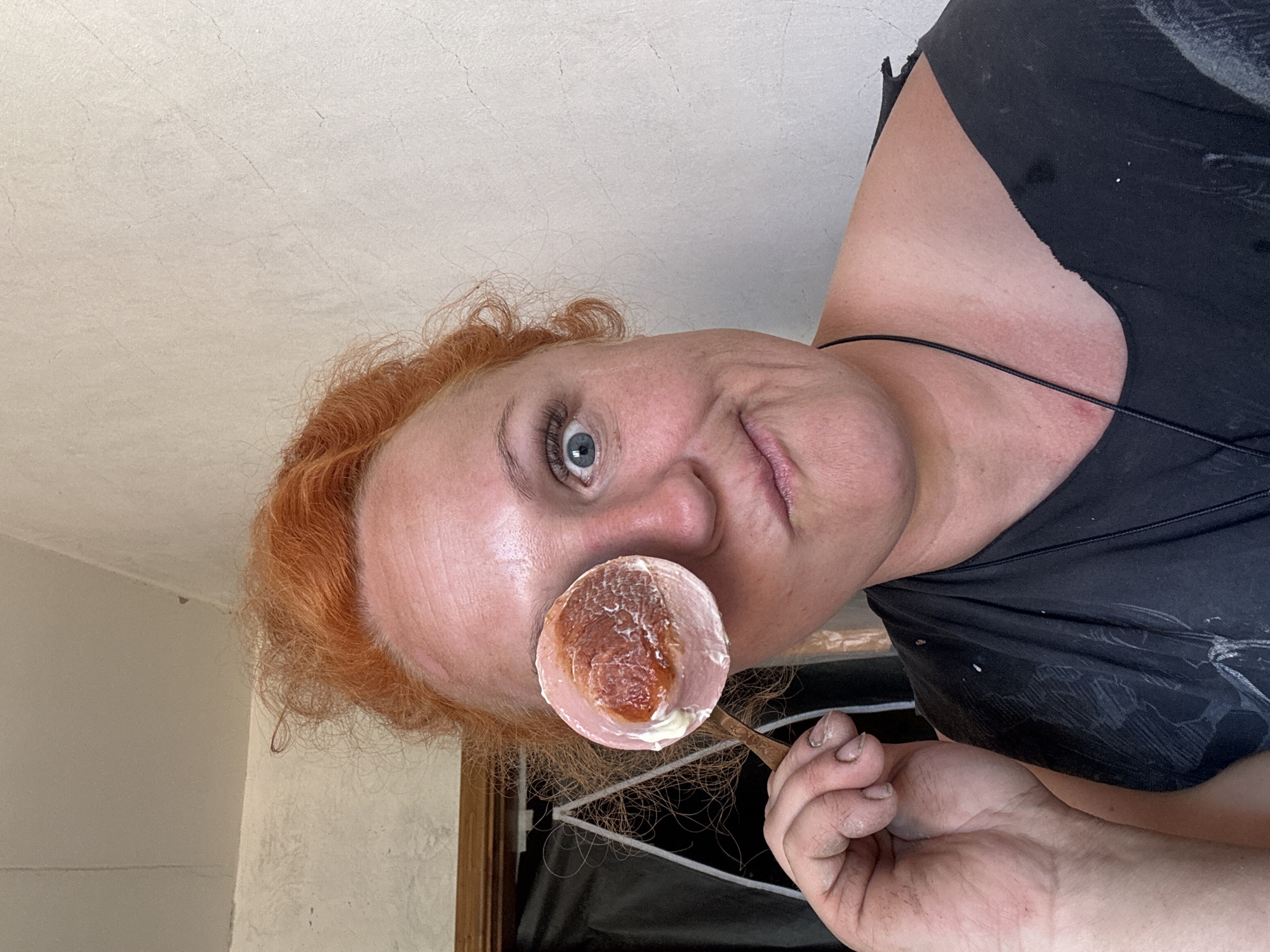 A woman with red curly hair and fair skin is holding a spoon with a pastry-like substance in front of her face. The woman has one visible blue eye. She is wearing a black shirt and a thin black necklace. Her hand is dirty. The background appears to be an interior setting with white walls and a dark object partially visible behind her head. The image is angled and may have been taken from a high vantage point. There is no definitive information in the image to confirm the location as Chaikovsky, Russia.