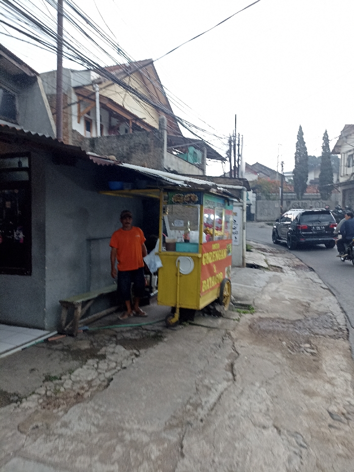 The image shows a street scene in Jatinangor, Indonesia. It appears to be a narrow, residential street with a small food cart parked on the sidewalk. There is one man standing next to the cart and he is wearing an orange shirt and blue shorts. The food cart is yellow with a red sign that says "Gorengan & Bakso" in Indonesian. There is a gray building to the left of the cart and a house in the background on the right. The street is paved with cracked concrete and there are power lines overhead. There is a dark-colored car parked in the street to the right of the food cart. There is a second, smaller, dark-colored car further down the road. A person is riding a motorcycle in the distance on the right side of the road. The scene is lit with a natural daylight, suggesting it is mid-morning or afternoon.