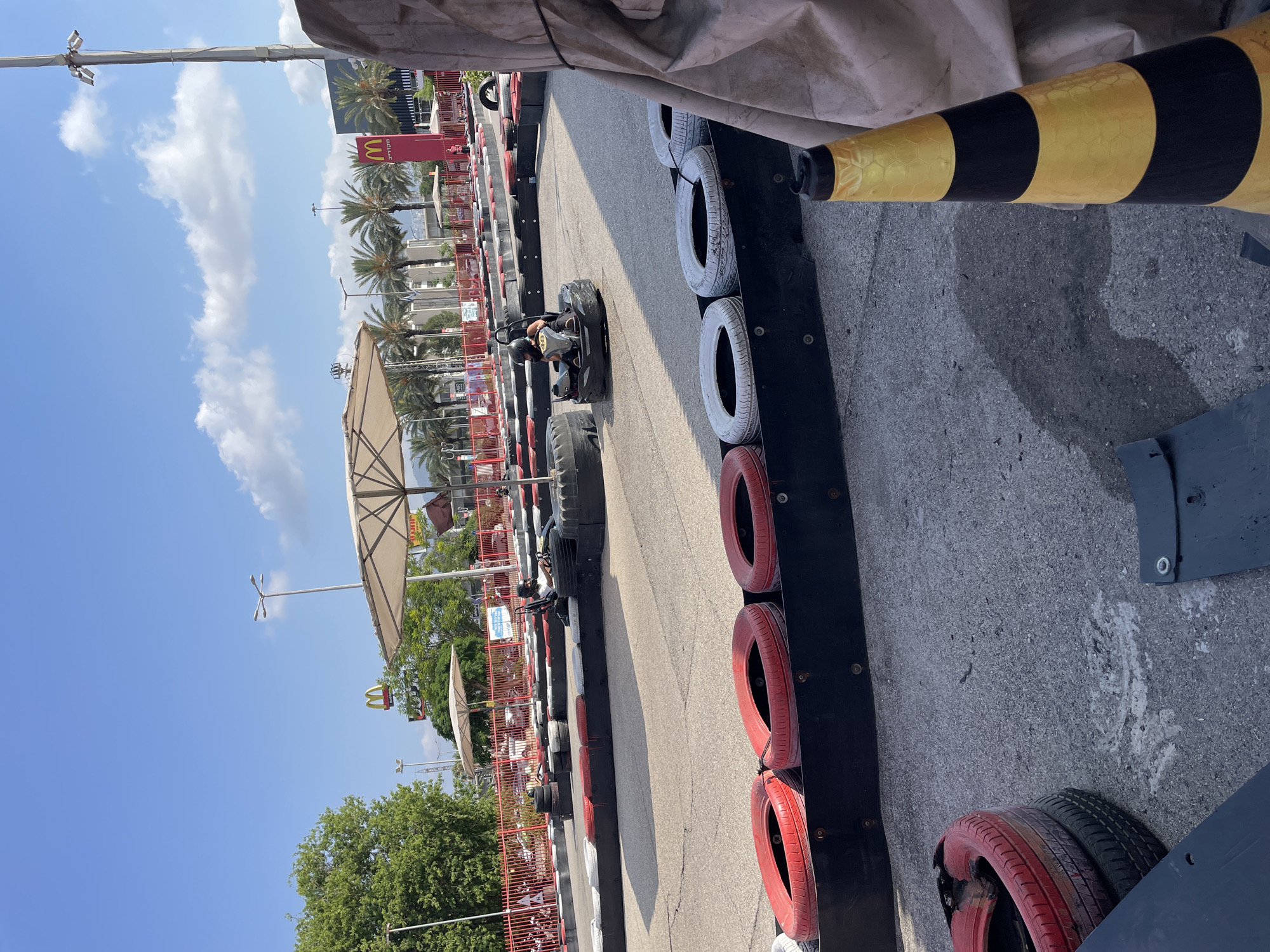 The image depicts a go-kart track with a person driving a go-kart in the foreground. The track is lined with tires for safety barriers, some of which are painted red. In the background, palm trees and buildings are visible, including McDonald's signs, suggesting an urban setting. The sky is blue with some clouds. The presence of McDonald's signs suggests this location is likely near a commercial area. Given the information that the location is Haifa, Israel, this is likely a go-karting facility situated within or near a shopping mall or entertainment complex in the city. The go-kart itself is black and low to the ground, and the driver is wearing a helmet. The overall impression is a recreational activity taking place on a sunny day.