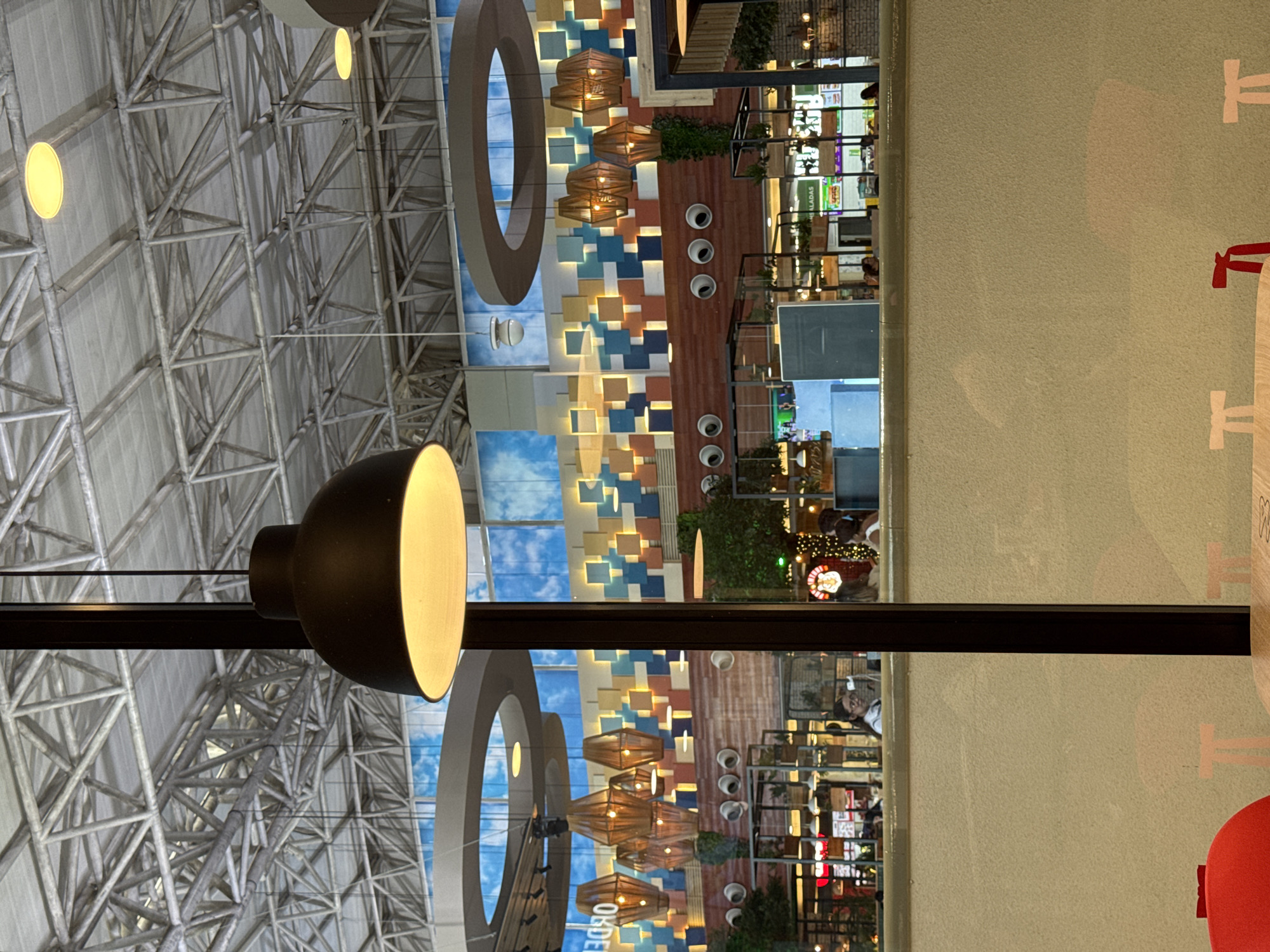 The image captures the interior of a large, modern commercial space, likely a shopping mall or a food court, in Porlamar, Venezuela. The perspective is looking upwards and across.
In the upper section, an intricate industrial-style ceiling is visible, characterized by a crisscrossing metal truss structure and several circular embedded lights. A prominent black pendant light with a warm yellow glow hangs in the foreground.
Below this, the scene transitions to a brightly lit dining or retail area. A striking decorative wall is featured, constructed from rectangular tiles in various shades of blue, brown, and beige, illuminated by hidden accent lights. Several woven, basket-like pendant lamps emit a warm glow in front of this wall. A large, suspended brown ring also serves as a decorative architectural element. Reflections of a blue sky with clouds are visible on surfaces, suggesting either skylights or reflective design features.
In the background, numerous people are seen, some seated at tables in a casual dining setting, others walking between establishments. The far right side of the image reveals a plain, light green wall adorned with stylized white and red painted graphics depicting tables and chairs, with a red seating cushion partially visible in the corner.
The environment appears modern and active. Visible text includes "SALADAS" on a sign in the background, likely advertising a food item, and "OBES" partially visible on another reflective surface. The time of day appears to be bright, with internal lighting complementing the ambient light.