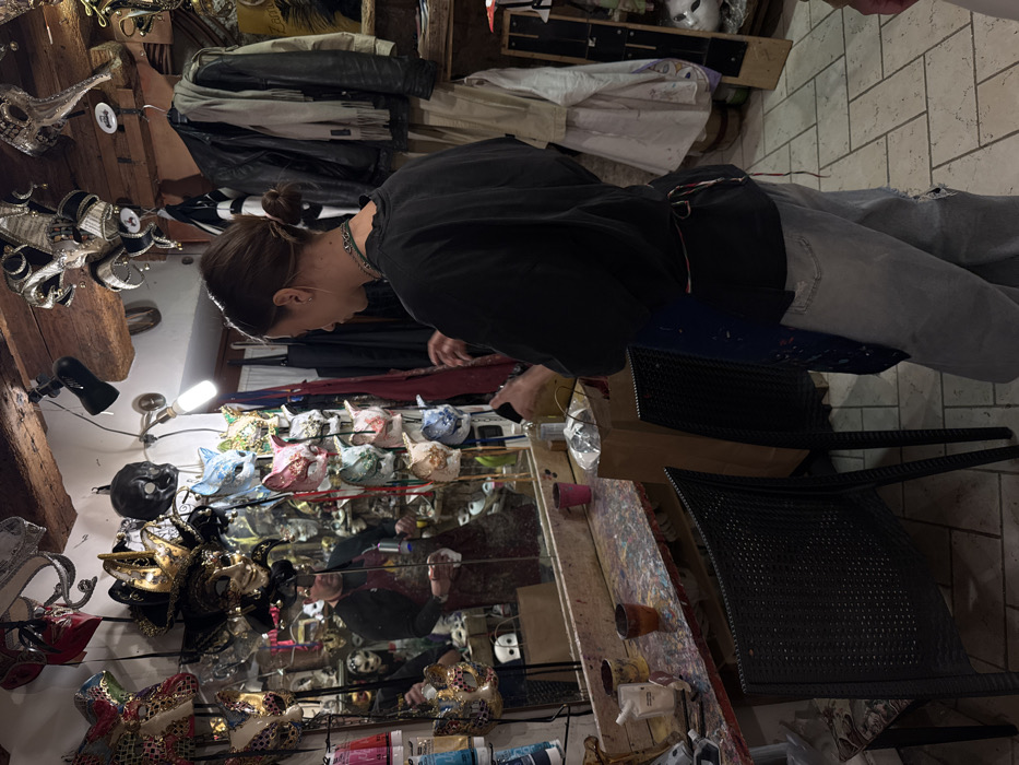 The image depicts the interior of an artisan mask workshop or retail shop in Venice, Italy. A young adult female with dark hair tied in a bun, wearing a dark t-shirt, light blue ripped jeans, and a dark blue apron, stands in the right foreground, facing left towards a paint-splattered wooden workbench. Her hands are positioned over a brown paper bag and a clear bottle. Reflected in a large mirror that forms the backsplash of the workbench, an adult male with a beard and a dark red apron is visible, holding a white mask in his left hand and a spray bottle in his right, actively working on the mask.
Hundreds of elaborate Venetian-style masks, in various colors, designs (including animalistic, jester, and traditional forms), and materials, are displayed on the walls and suspended from the wooden-beamed ceiling. The workbench surface is heavily stained with multiple colors of paint, and various art supplies, including paint tubes and small cups, are present. Two dark-colored chairs are positioned in front of the workbench. The room is illuminated by artificial light sources, including a bare bulb and a spotlight. Clothes, including jackets and scarves, hang on racks on the right side of the room. The scene indicates an active artistic environment dedicated to mask creation and display.