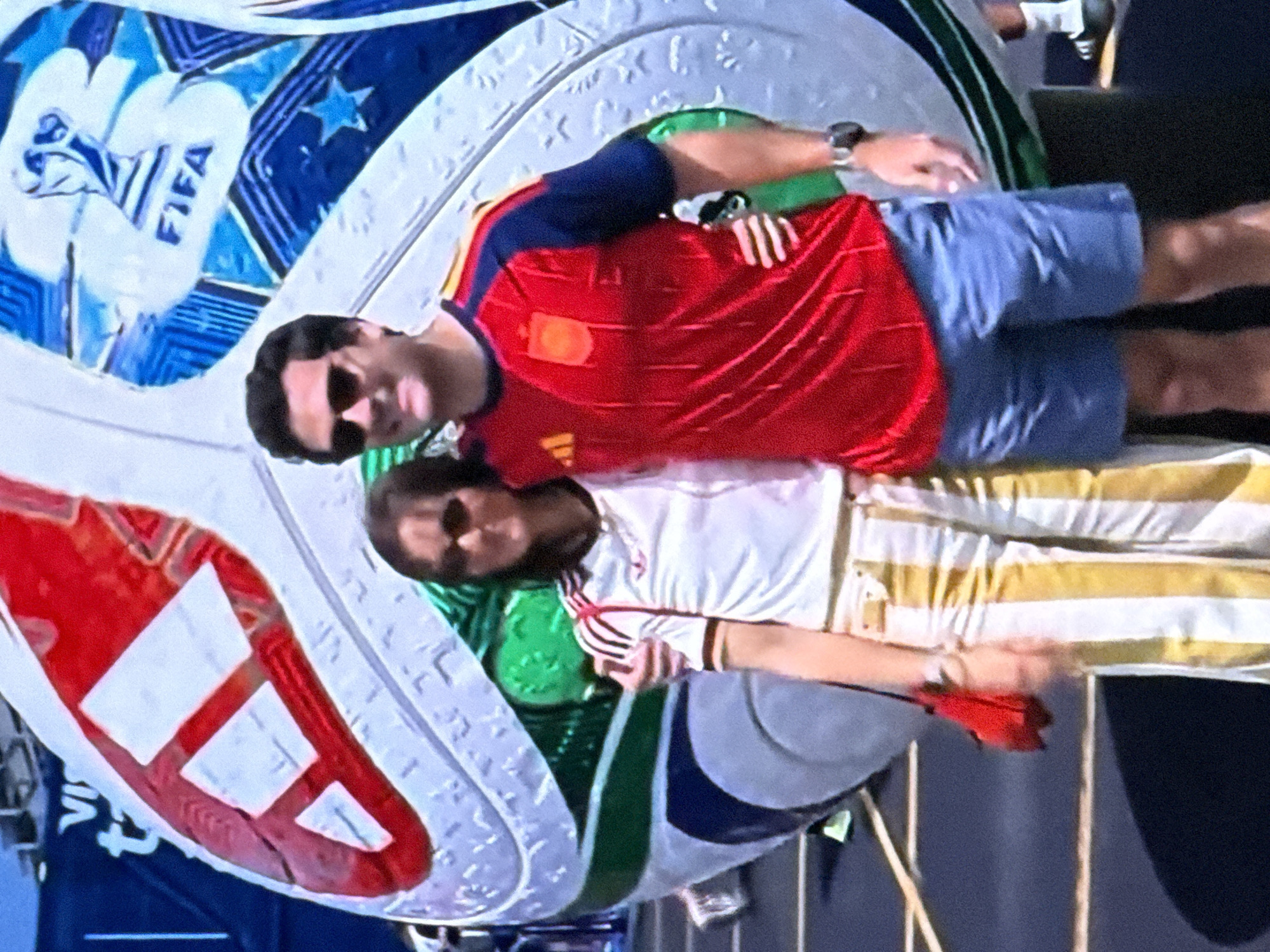 In Malmyzh, Russia, a couple poses in front of a giant soccer ball prop at an outdoor event. The man wears a red and blue soccer jersey with the Spanish national team crest and blue shorts. The woman is dressed in a white Adidas shirt with red stripes and yellow and white striped pants, carrying a red purse. The soccer ball, branded with FIFA and Adidas logos, dominates the background. The ground is paved and marked with white lines. The lighting suggests it is daytime.