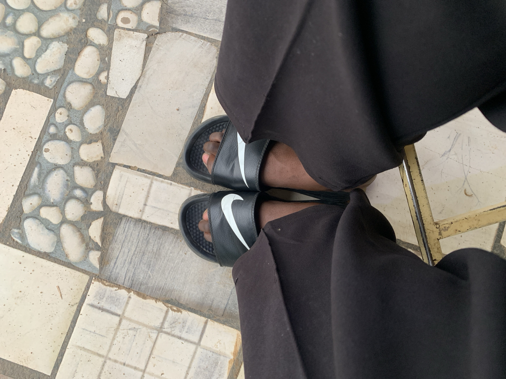 A person wearing black Nike sandals and baggy black pants is sitting in a chair. The chair has a metal frame with chipped yellow paint. The person is seated on a tiled surface, which includes sections of mosaic tiles, arranged with white pebbles. The image appears to be taken from an overhead perspective. The location is New Stadium, Nigeria.