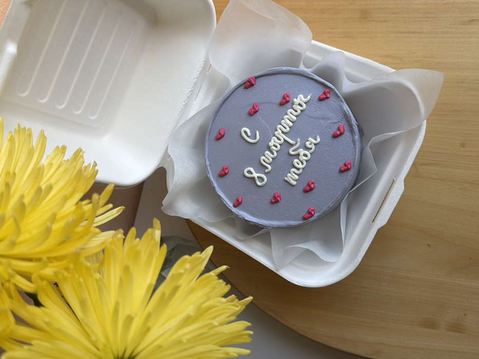 A small, circular cake, frosted gray and adorned with white piped Cyrillic text reading "С 8 марта тебя" (Happy March 8th to you), is presented within an open white disposable container. Scattered across the cake's surface are approximately a dozen small, red, heart-shaped icing decorations. The cake rests on translucent white tissue paper inside the container. The upper portion of the container's ribbed lid is visible in the background, open. The objects are situated on a light brown wooden surface. In the upper-left of the frame, a cluster of bright yellow flowers with thin petals, consistent with chrysanthemums, is partially visible. The scene is located indoors, in Tel-Aviv, Israel.