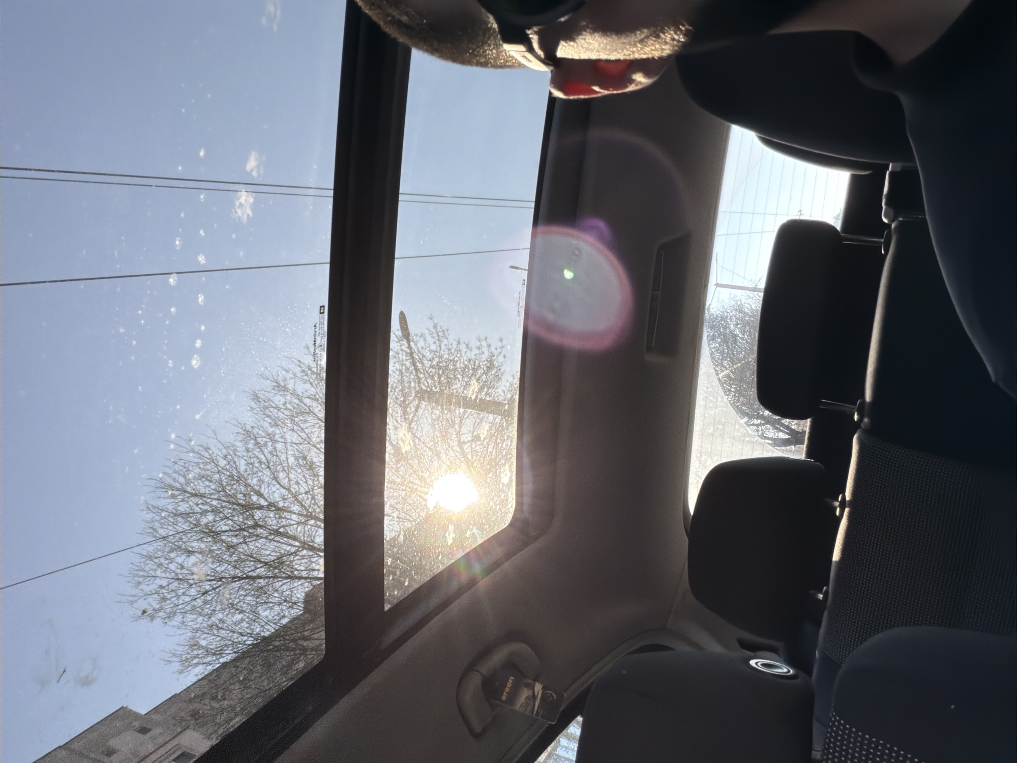The image shows a view from inside a vehicle, likely a car, through an open sunroof. The sky is visible, with a bright sun causing lens flare. There is a tree with bare branches outside the window. Buildings can be seen in the background. The interior of the car includes seats with dark upholstery, headrests, and parts of the car frame and ceiling. Given the context of Sofia, Bulgaria, the architecture in the background is likely typical of the city. There is a person visible in the top part of the image.
