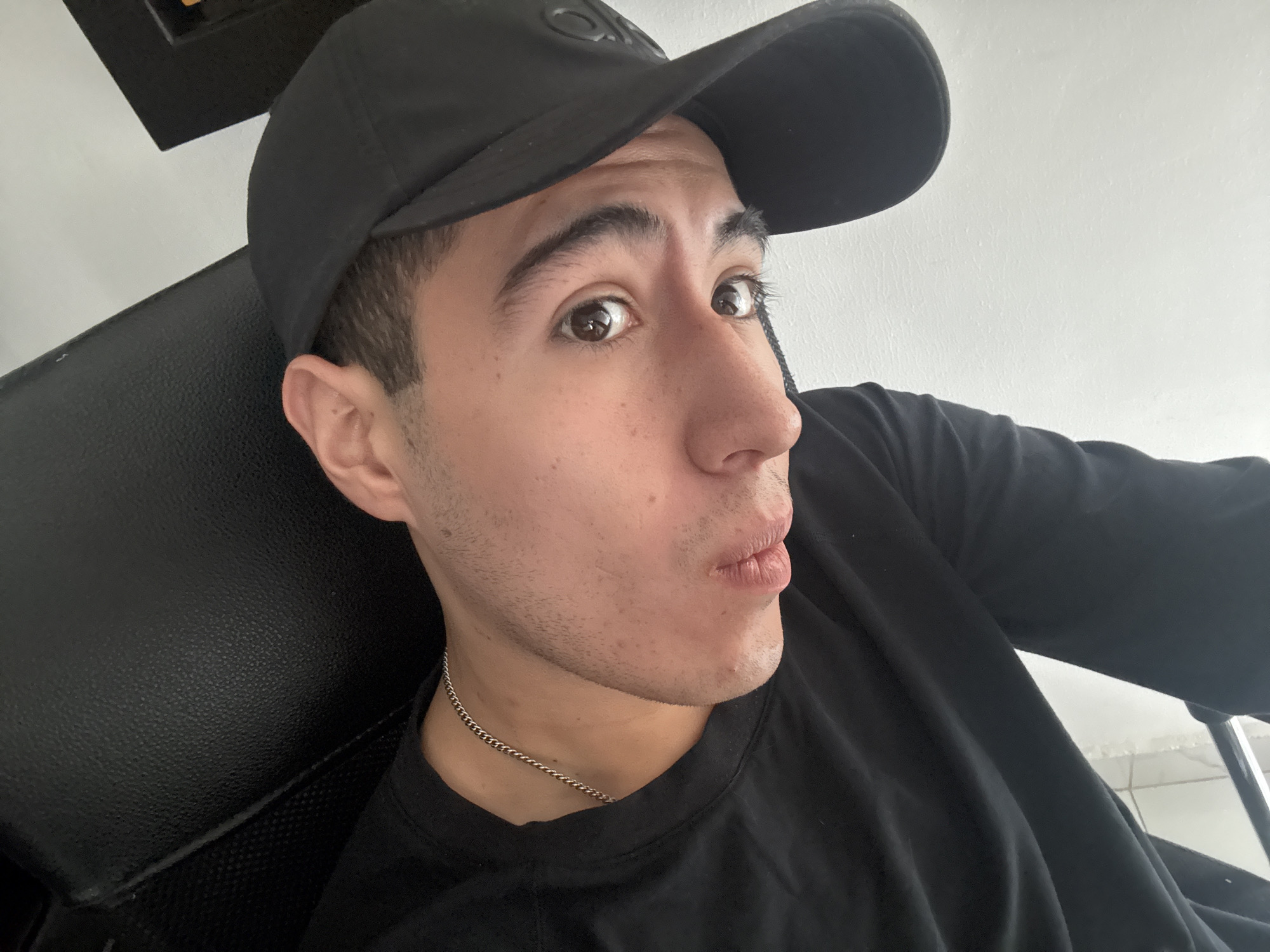 A young man is seated in a black chair, wearing a black T-shirt, a silver chain necklace, and a black baseball cap. He is looking at the camera with a slightly puckered expression. The background appears to be a white wall with a dark-colored object affixed to it, possibly a picture frame or some other decorative element. The location is not evident from the image. There is no way to determine if the image was taken in San Bernardino Tlaxcalancingo, Mexico.