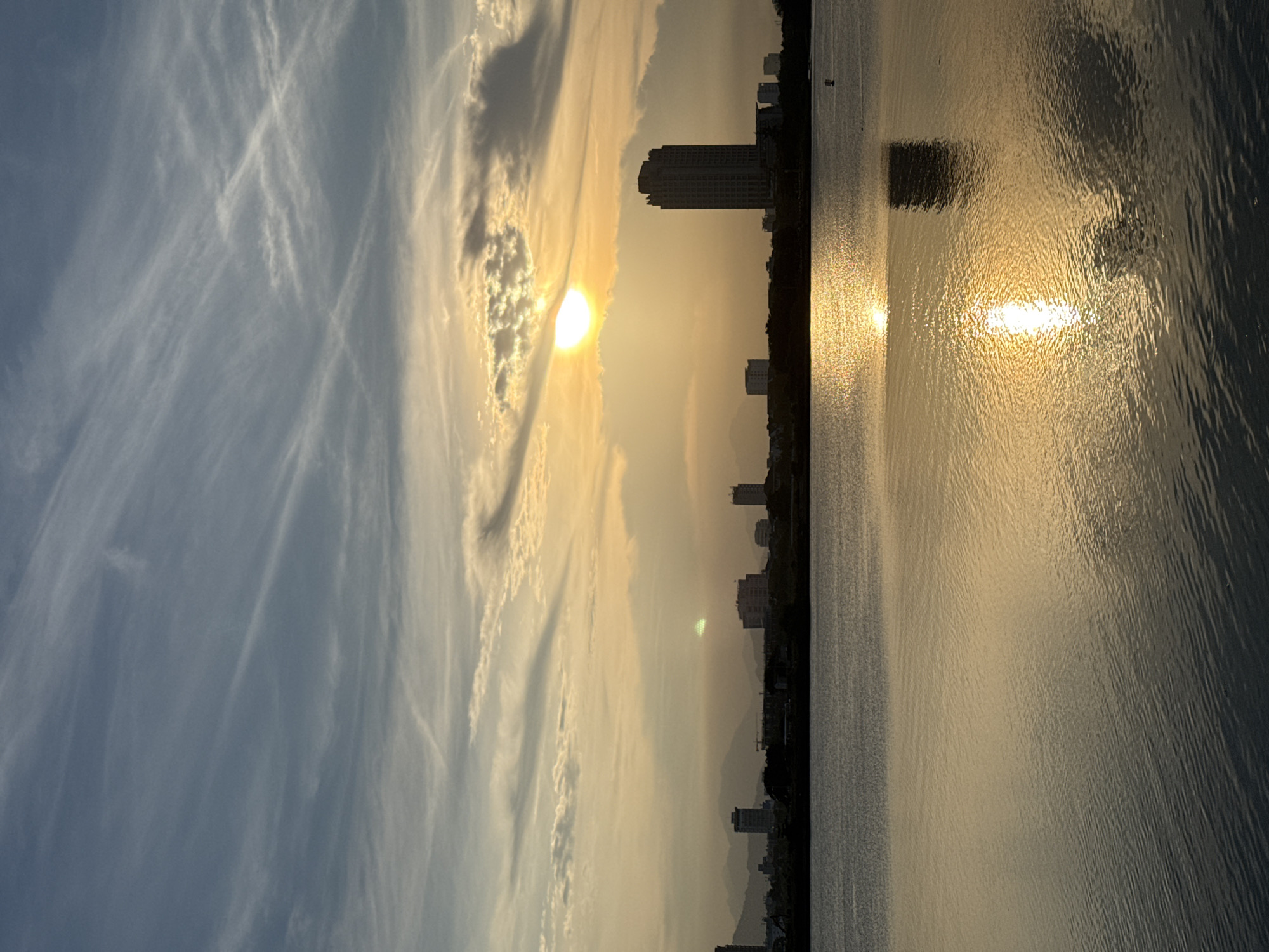 This image captures a sunset over the Han River in Da Nang, Vietnam. The sun, partially obscured by clouds, casts a golden glow across the sky and reflects vibrantly on the water's surface. Silhouetted against the colorful sky is the Da Nang skyline, featuring several tall buildings, including the distinctive Đà Nẵng Administration Center tower. The river forms the foreground, with gentle ripples disturbing the water's mirror-like surface. The horizon line separates the water from the city and distant mountains, creating a layered composition. The overall atmosphere is serene and picturesque, indicative of a peaceful evening in the city.