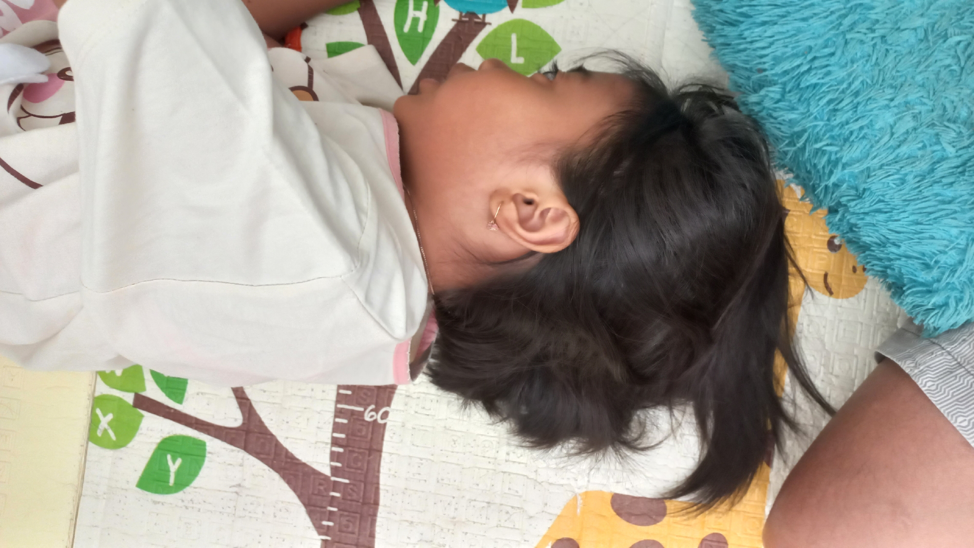 A young child, lying on their side with their head facing right, rests on a mat in what appears to be a residential setting in Jetis Indah, Indonesia. The child is wearing a white t-shirt with pink trim and grey striped shorts. Their dark hair is spread around their head, and a small, pink earring is visible in their right ear. A blue, textured blanket or pillow is positioned to the child's right. The mat beneath the child features a colorful pattern with trees, leaves, and what seem to be cartoon animal faces. The overall environment suggests a casual, indoor space, possibly a play area or bedroom.