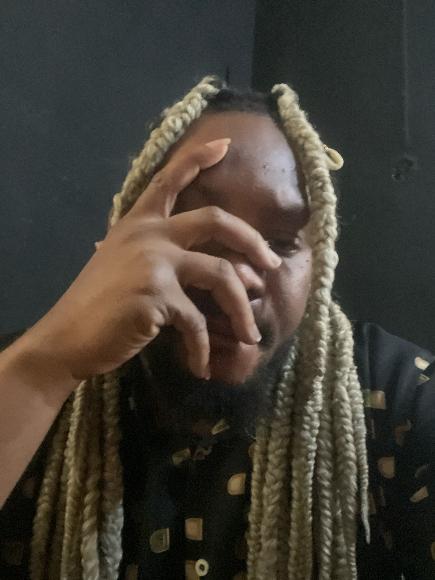 The image depicts a close-up of a person with light-colored braids, with their hand resting on their forehead, obscuring part of their face. The background is a plain, dark surface, possibly a wall. The individual is wearing a black shirt with a pattern of light-colored squares and circles. There is no visible indication of the city of Jos, Nigeria, or any specific environmental context that would suggest this location. The lighting appears to be dim, creating shadows on the person's face and hand.