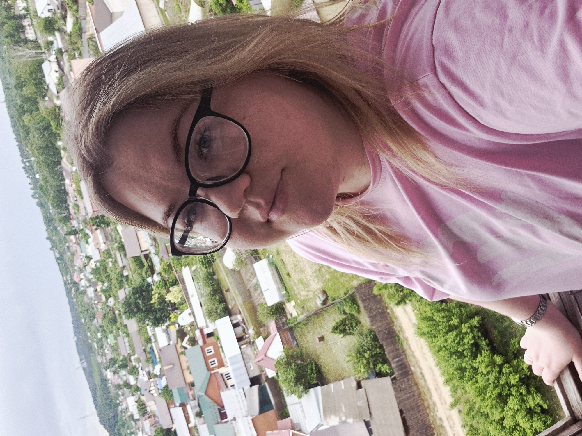 A fair-skinned woman with blonde hair and black-framed glasses poses for a selfie. She's wearing a pink T-shirt and a silver watch on her left wrist, and her hand grips a wooden railing. The background reveals a view of the city of Chaikovsky, Russia, with houses and trees visible from an elevated perspective.