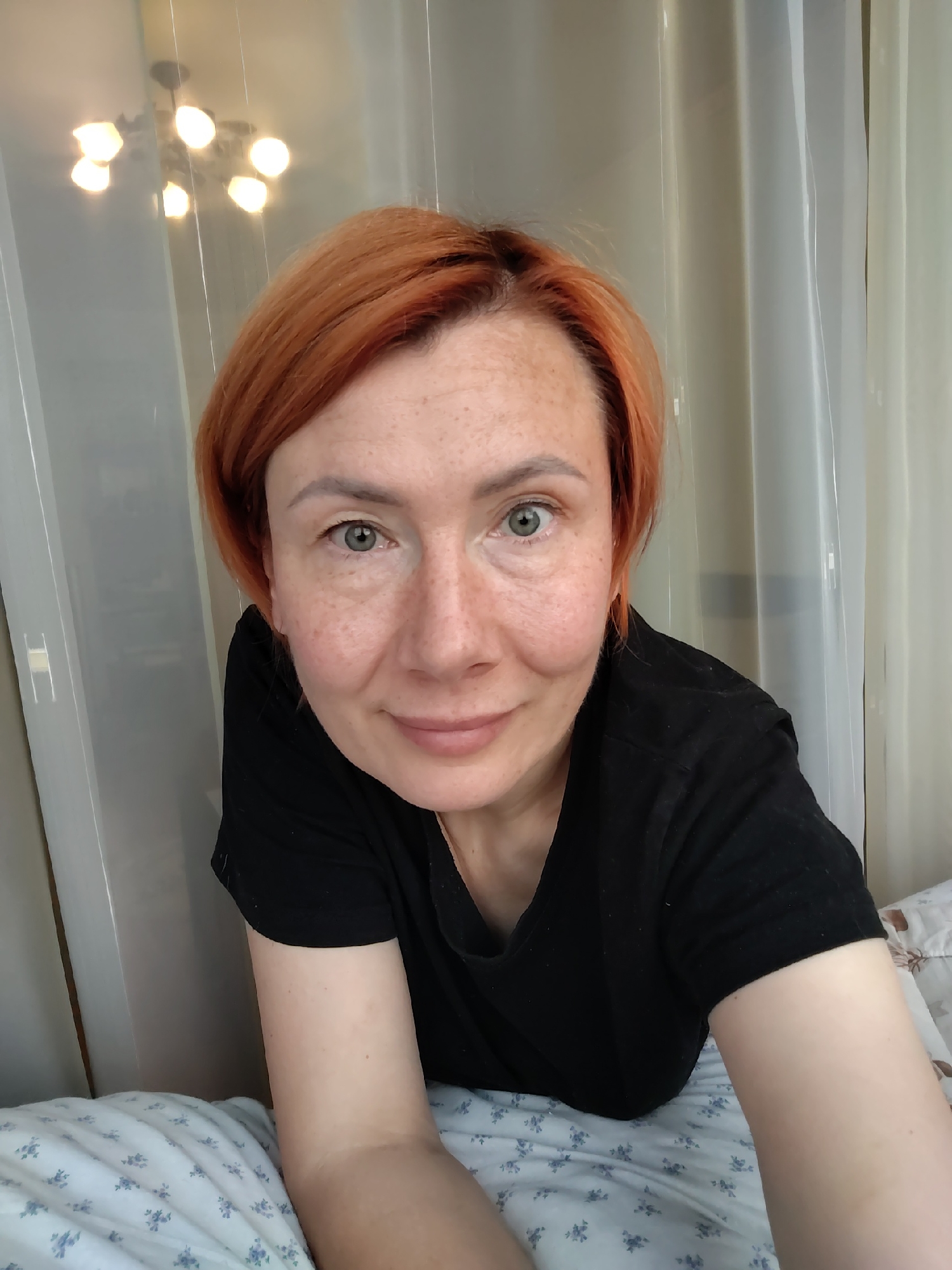A medium shot, waist-up, selfie shows a fair-skinned Caucasian woman with short, reddish-orange hair and freckles on her face. She is wearing a black t-shirt and is lying down on a bed with a white and blue floral patterned duvet. Her face is turned towards the camera, and she is looking directly at the viewer with a slight smile. Her green eyes are open, and her eyebrows are light brown. Her arms are resting on the bed. The background consists of sheer white curtains and a chandelier with multiple lights visible in a reflection. The lighting appears to be natural, coming from a window. The scene suggests an indoor setting, likely a bedroom. The provided image is not sufficient to determine the location in the city of Konakovo, Russia.