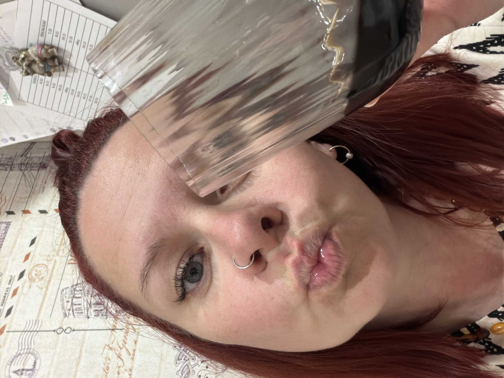 A young woman with red hair and fair skin is lying on her back. She is holding a clear plastic cup, filled with a dark liquid, near her face. Her lips are pursed in a kiss. She is wearing a nose ring in her left nostril and a small hoop earring in her left ear. The background is a surface with a printed pattern resembling vintage postcards. A document with printed text and a small toy are visible in the upper left corner. The location is likely Chaikovsky, Russia.