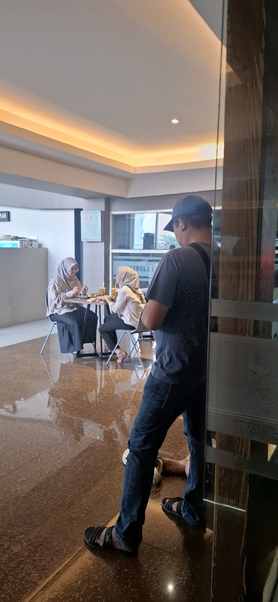In the city of Surakarta, Indonesia, inside what appears to be a cafe or restaurant with a reflective tiled floor and modern ceiling lighting, two women wearing hijabs are seated at a small round table, engaged in eating or conversing. One woman is seated on the left, facing the table, while the other is on the right, also facing the table. A man wearing a dark t-shirt, dark jeans, a dark baseball cap, and dark sandals stands in the foreground, partially obscured by a glass partition. He is facing away from the camera, looking towards the seated women. The environment is indoors, with a bright ceiling light strip. Visible on the wall behind the women is a white electrical box with colored indicator lights. The overall scene suggests a casual dining or meeting setting.