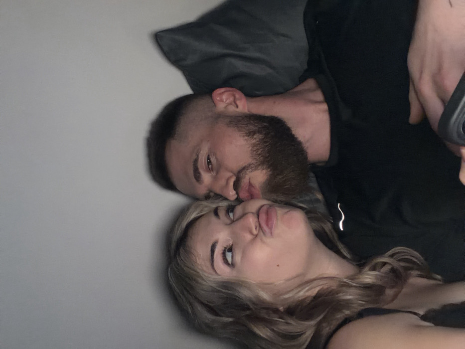 A man with a beard and a woman with long blonde hair are lying close to each other, angled diagonally in the frame. The man is wearing a black top, and the woman appears to be wearing a dark-colored top as well. They are both pouting their lips and looking at the camera. A dark pillow is visible behind the man's head, and the background is a plain gray wall. The photo seems to be taken with a camera or phone held at a slight angle.