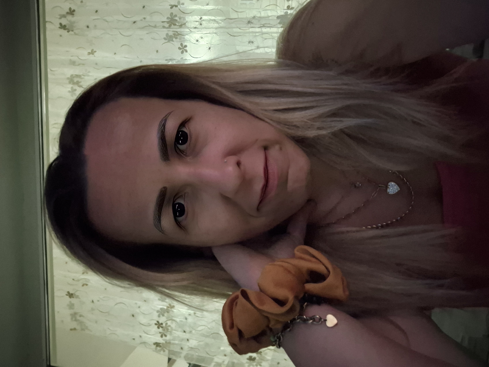 A medium close-up selfie of a white woman with blonde hair and light brown eyes is shown. She is looking directly at the camera. Her left hand is resting on her cheek, and she is wearing a scrunchie on her wrist. She is also wearing a necklace with a heart-shaped pendant. The background appears to be a room with light-colored curtains.
