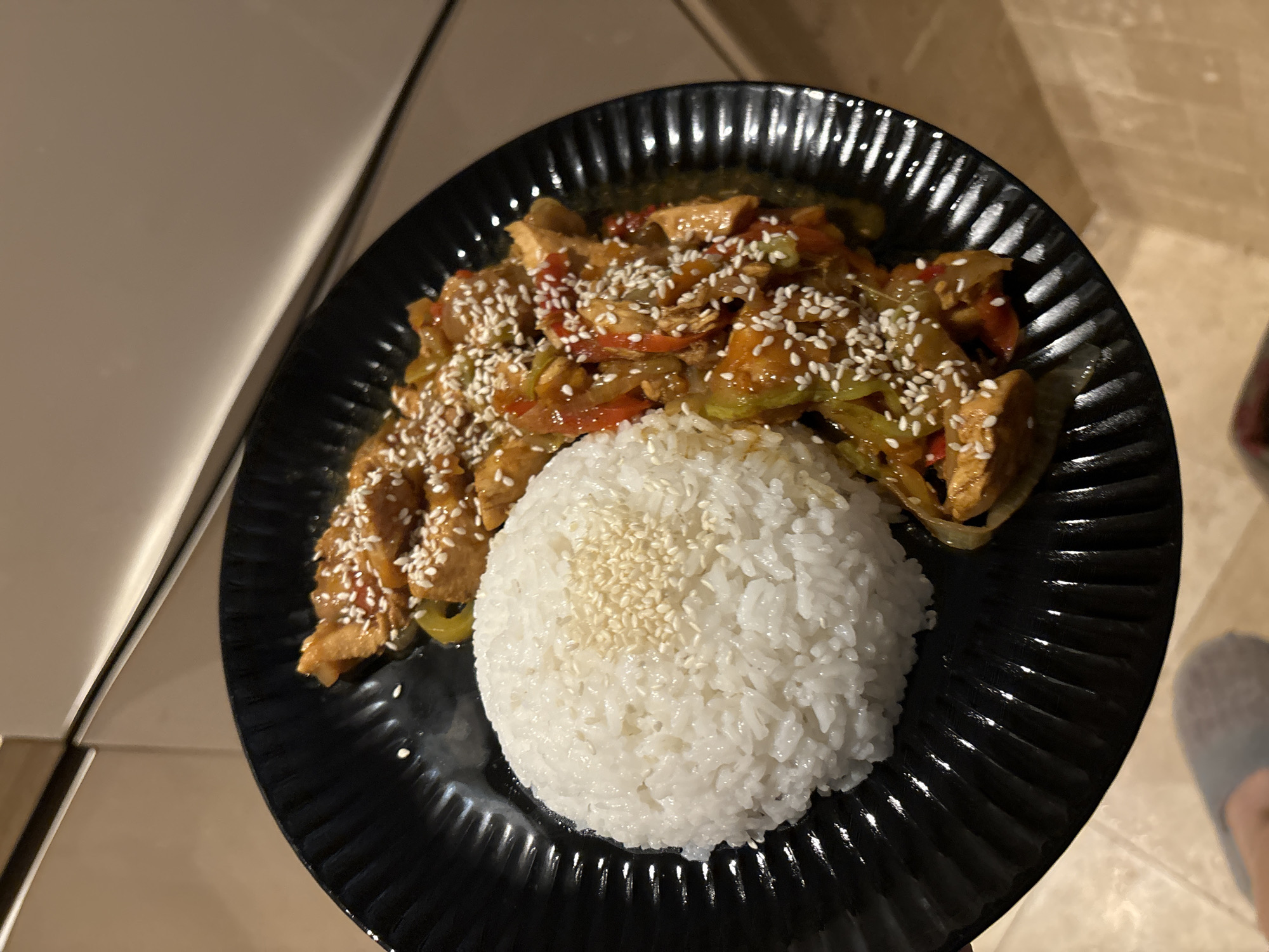 A top-down, close-up shot shows a black plate containing cooked chicken, vegetables, rice, and sesame seeds. The chicken and vegetables, which include peppers and onions, are coated in a dark sauce and sprinkled with sesame seeds. A large mound of white rice, also sprinkled with sesame seeds, sits adjacent to the chicken and vegetables on the plate. The plate has a ribbed pattern. The plate rests on a surface that appears to be a countertop. Part of a piece of furniture is visible in the upper left corner of the image. A foot in a slipper is visible in the lower right corner. The photo was not taken in Budva, Montenegro.