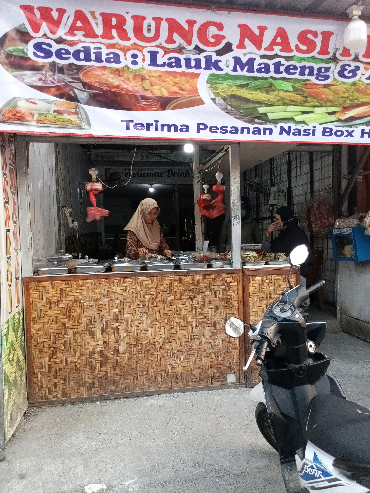 A street-level, medium shot captures a food stall named "WARUNG NASI" (Rice Stall) in Karangsatria, Indonesia. The stall features a prominent banner with red and blue lettering announcing its offerings. Inside, two women are visible; one in a light brown hijab is serving food from various metal containers, while another in a dark hijab is seated. The stall's counter is decorated with a woven bamboo pattern. In the foreground, a black and white motorbike is parked on the concrete ground, partially obscuring the stall. The environment appears to be an outdoor setting, typical of a local marketplace.