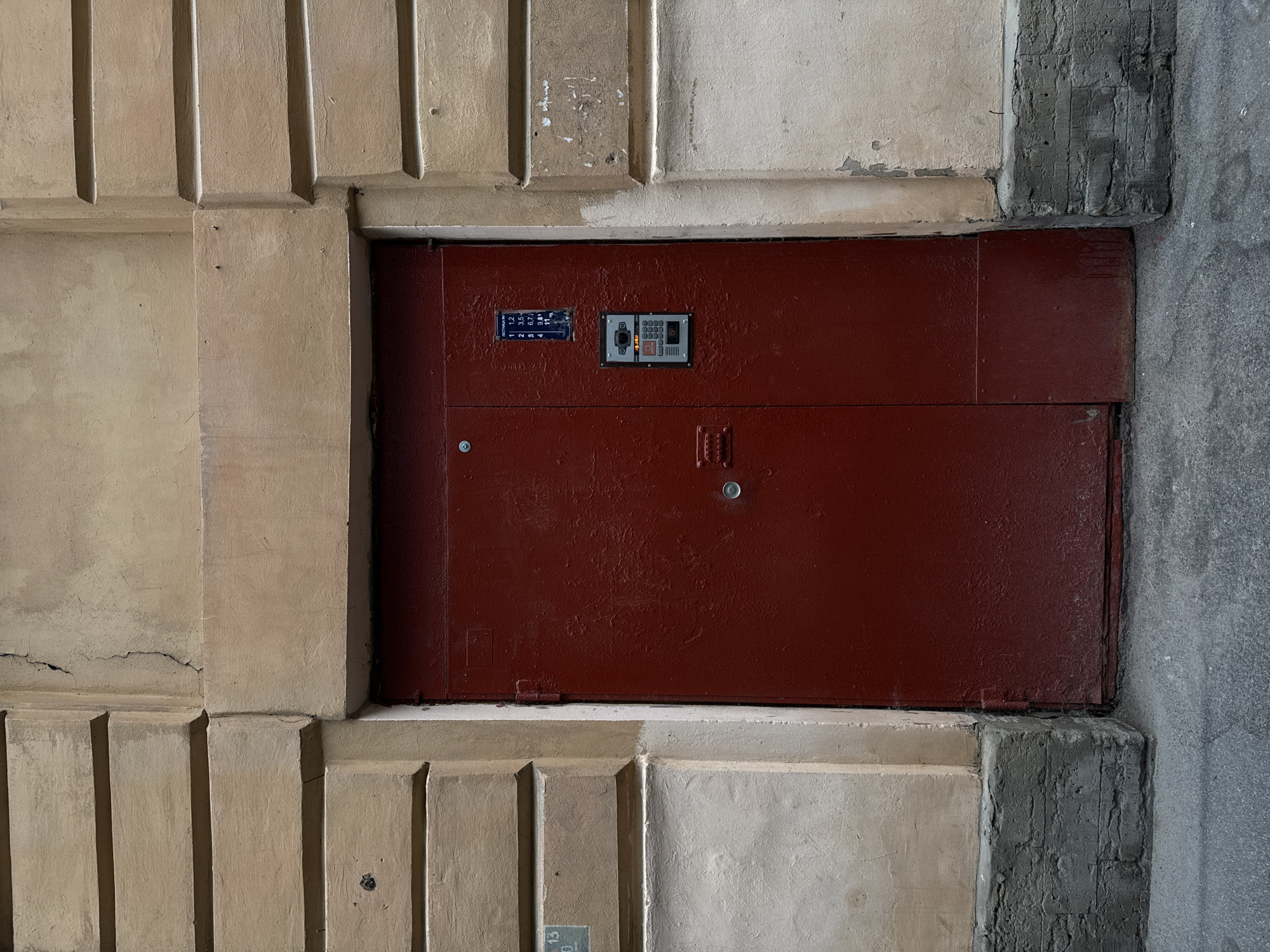 The image shows a weathered, dark red metal door in a light beige building facade in Saint Petersburg, Russia. The door has a metal interphone system and a keypad attached to its surface. The building's exterior is characterized by horizontal segmented stone blocks above and a rough, off-white plaster below. The foundation of the building is a dark, textured concrete block. The foreground displays a gray asphalt surface. The overall impression is one of a functional, urban entrance with signs of age and wear.