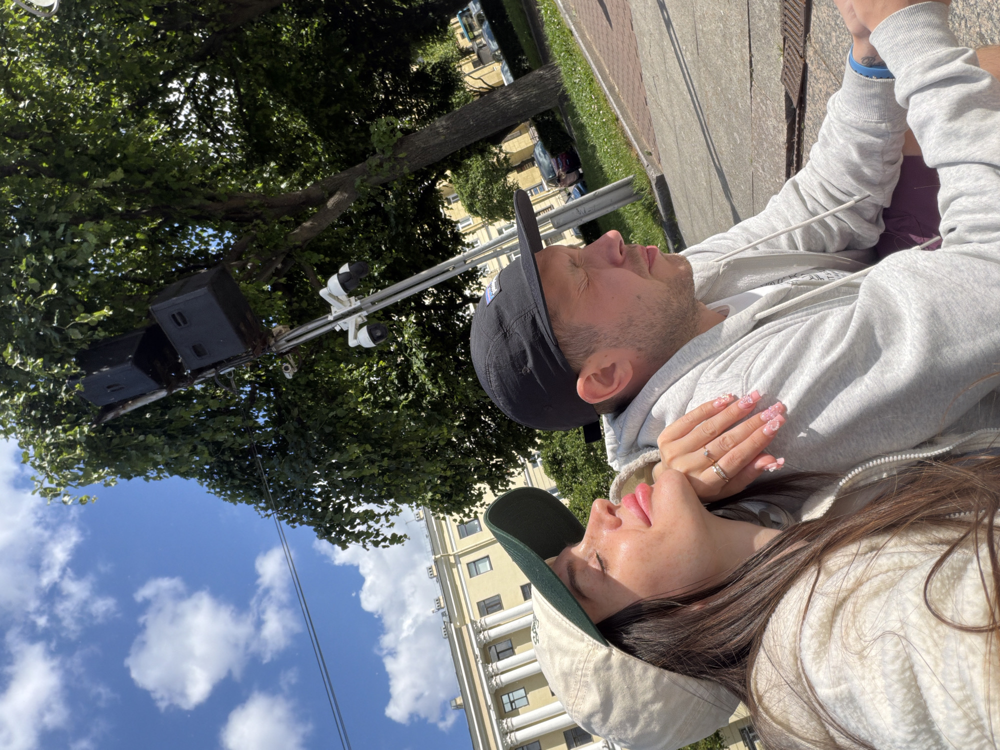 A medium shot shows a couple sitting outdoors, with a building and trees in the background. The woman, on the left, is wearing a beige baseball cap and a white puffer jacket. She has her eyes closed and her head tilted back, with her chin resting on her hand. The man, on the right, is wearing a black baseball cap and a gray hoodie, with his eyes closed. They are both looking towards the sky. In the background, a light yellow building with white columns and windows is visible. Trees with green leaves are also present. The scene appears to be a sunny day, with a blue sky and white clouds. The location is identified as Saint Petersburg, Russia, with a prominent street lamp and what appears to be security cameras visible.