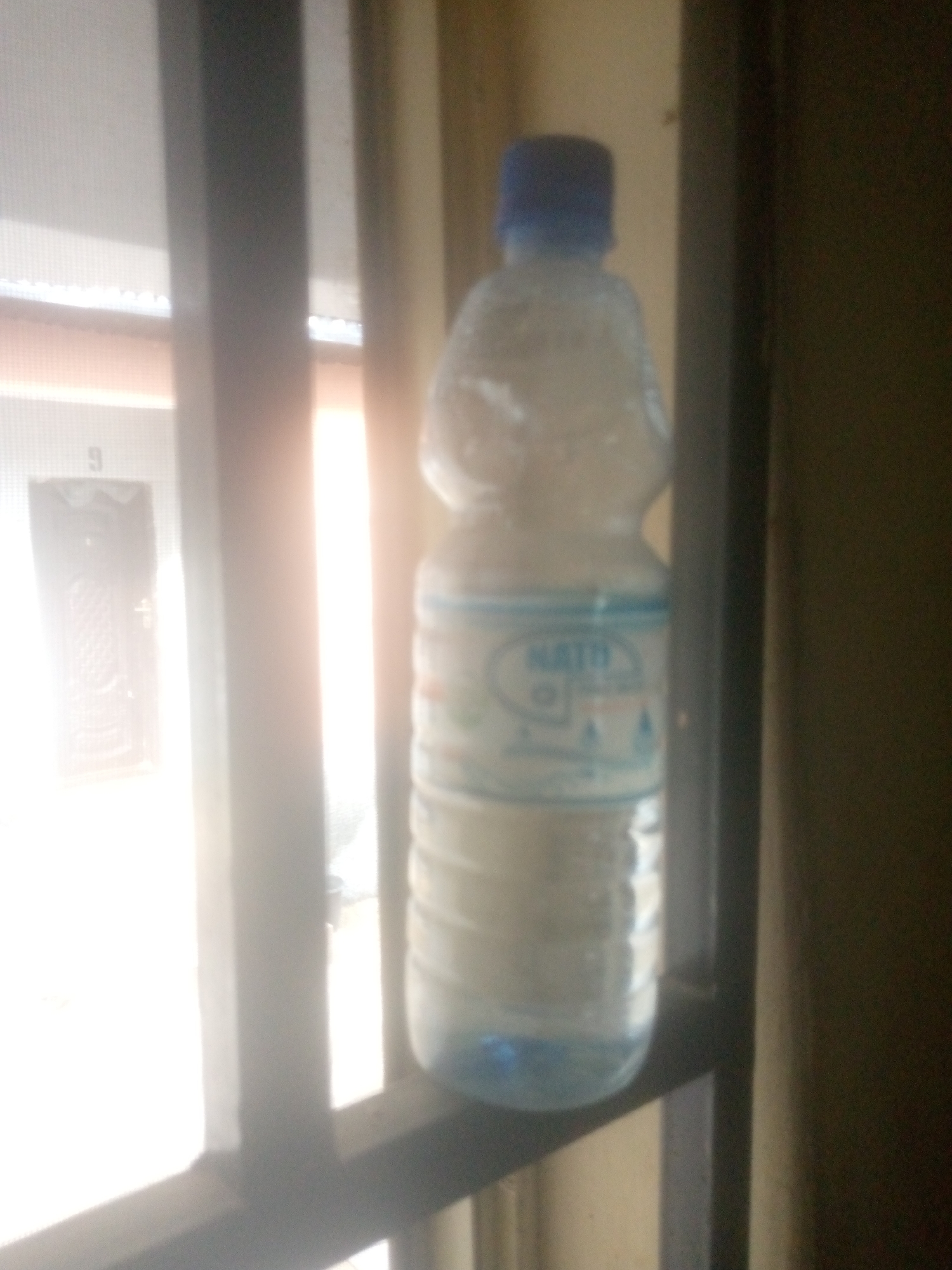 A plastic bottle of water is placed on a window sill. The bottle is partially filled with water and has a blue cap. The label on the bottle is partially visible and appears to say "MATI". The background is blurry and shows a building with a door numbered "9". The scene is brightly lit, suggesting it is daytime. The overall image quality is poor, with low resolution and poor focus. The location is unknown.
