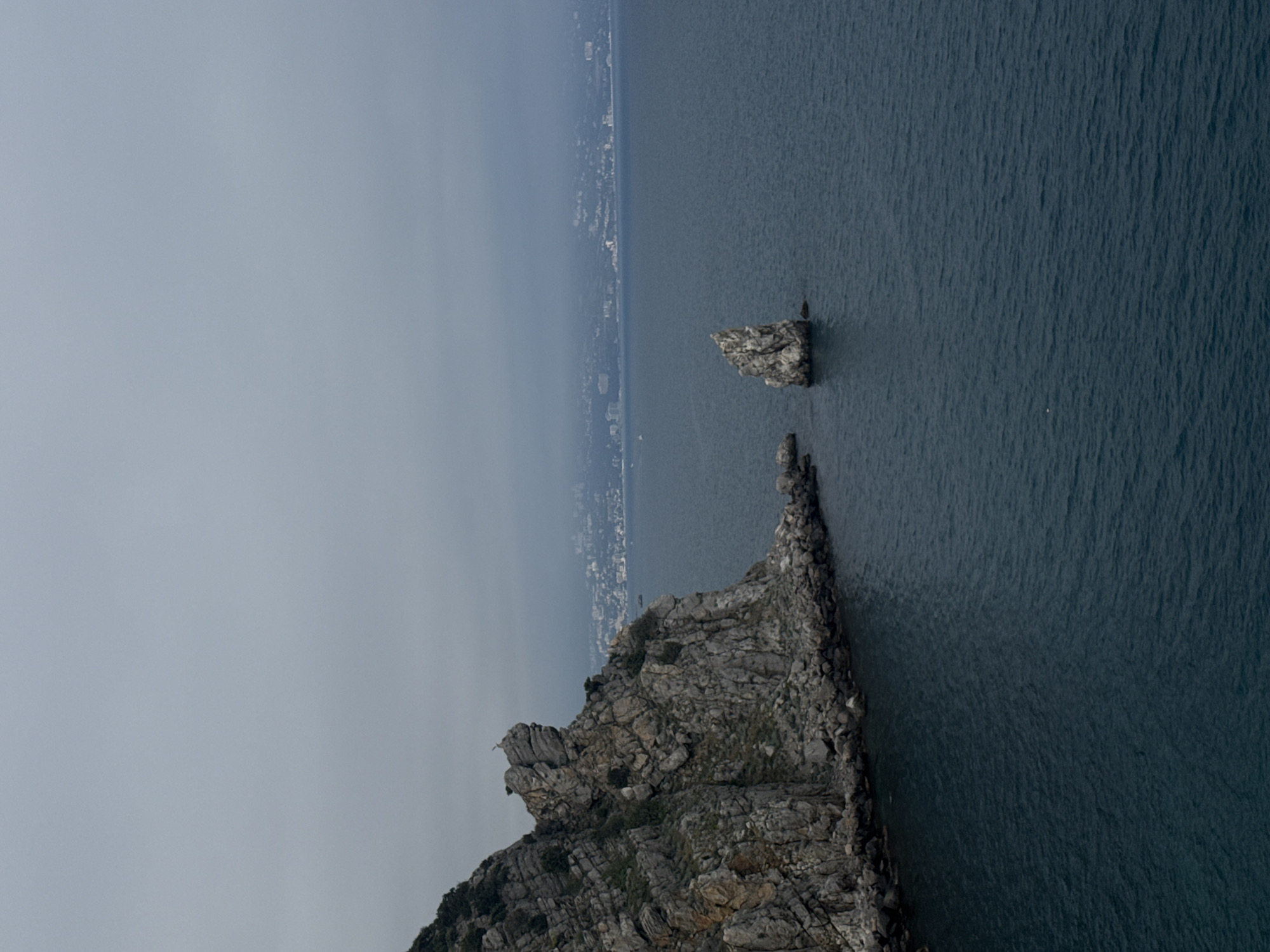 The image features a rocky cliff formation jutting out into the sea, with a smaller, isolated rock formation nearby. The sea is relatively calm and dark blue. Along the horizon, a coastline with a dense concentration of buildings suggests a city or town. The sky is overcast and gray, with a visible haze. The photograph appears to be taken from an elevated position, giving a bird's-eye view of the landscape. Based on the provided information, the location is likely in or near Гаспринский поселковый совет, Ukraine.