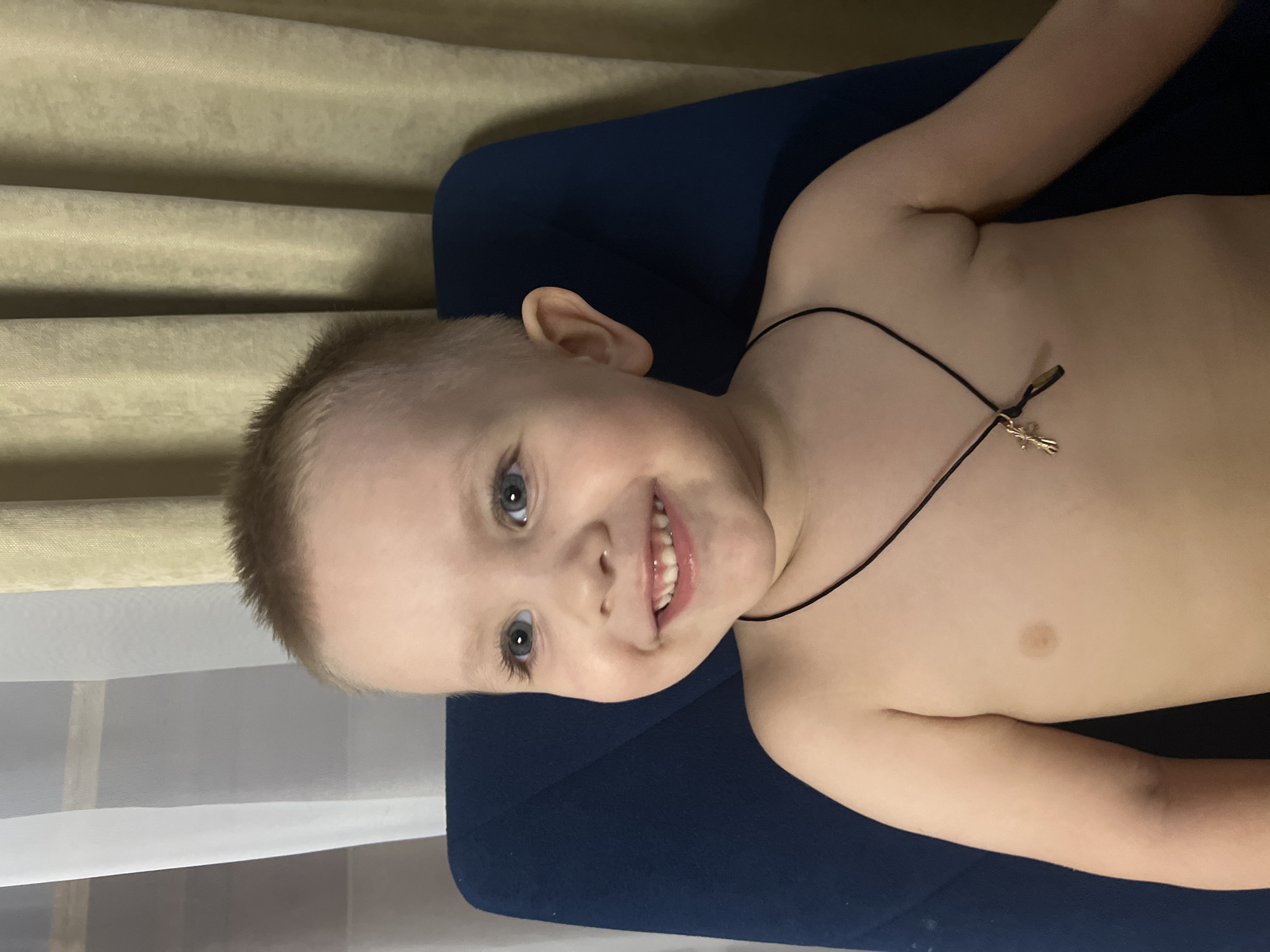 A shirtless child, likely a boy, is lying on a dark blue surface. He has short, light blonde hair, fair skin, and blue eyes. He is smiling, revealing his teeth. He is wearing a thin black necklace with a small silver cross pendant. The background includes sheer white curtains and a light brown curtain. The image was taken indoors. Location: Unknown, but requested location is Марковский, Russia.