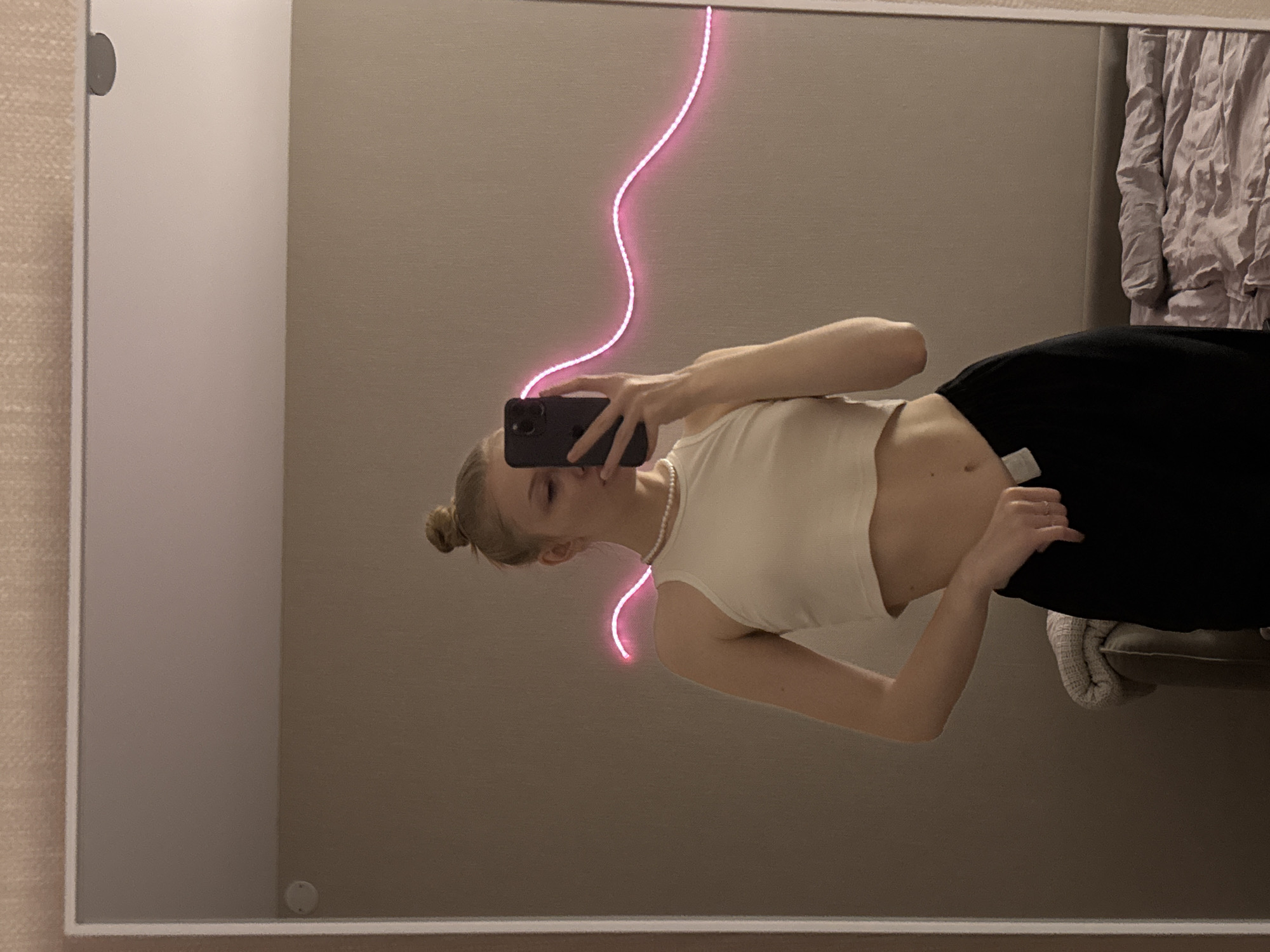 A young female is captured in a mirror selfie within an indoor setting. She has light-colored hair styled in a bun on top of her head. Her face is partially obscured by a dark-colored smartphone, held in her right hand, which she uses to take the photo. She wears an off-white sleeveless crop top, exposing her midriff, and dark-colored pants. Her left hand is positioned on the waistband of her pants, revealing a white tag. A single-strand pearl necklace adorns her neck, and at least one ring is visible on her left hand.
The reflection reveals a room with light beige walls, some featuring a textured wallpaper pattern in the upper section. A bright, wavy pink LED strip light is affixed to the wall behind her right shoulder, extending horizontally. In the background to the right, an unmade bed with light purple/lavender crumpled bedding and a neutral headboard is visible. To the left, an armrest of a piece of furniture, possibly a chair or sofa, is partially seen, draped with a rolled or folded light-colored blanket. The mirror itself has a thin, white frame. Two circular, flush-mounted fixtures (possibly lights or sensors) are visible on the upper wall/ceiling area within the reflection.