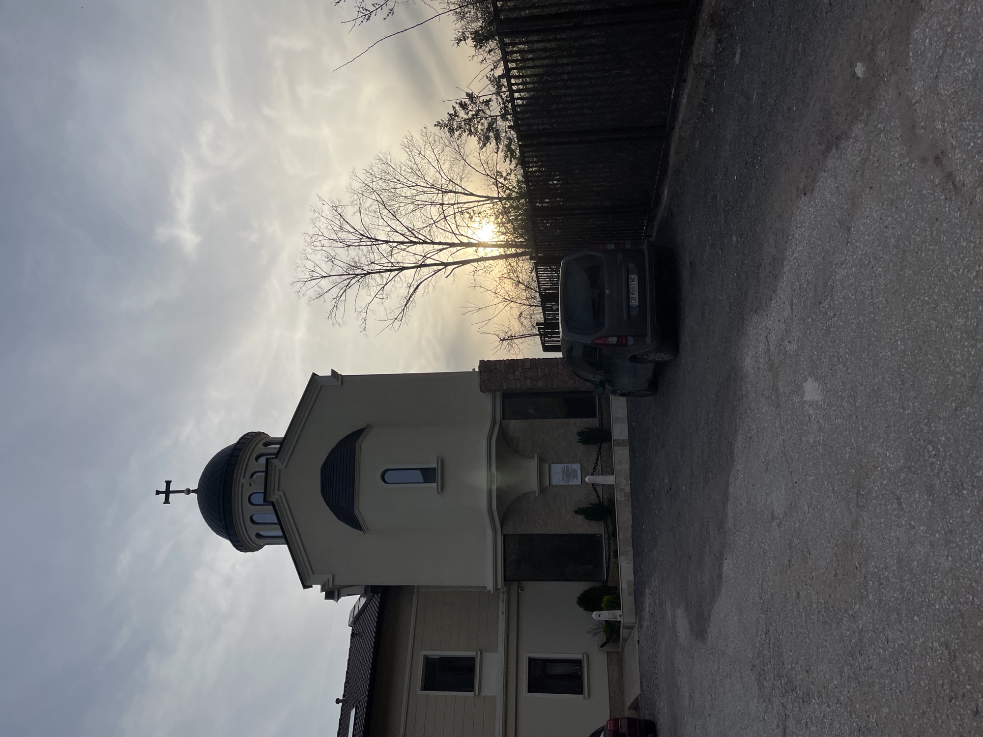 The image depicts a light-colored church building with a dark dome and a cross on top. The church is located next to a paved road, and a dark-colored van is parked on the side. A fence runs along the edge of the road. Bare trees stand between the church and the fence. The sky is overcast, with the sun shining through the clouds. The vehicle licence plate reads "CB 4551 MC". The location is Sofia, Bulgaria.