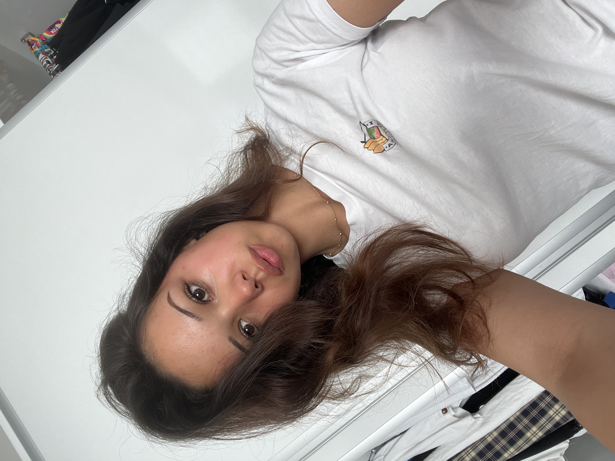 A young woman is taking a selfie in a room with white walls and a closet visible in the background. She has long, dark brown hair that is slightly tousled, framing her face. She is wearing a white t-shirt with a small, colorful patch on the left side of her chest. The patch appears to depict a stylized food item, possibly a sandwich or some sort of layered pastry, with an orange and brown color scheme. She has a delicate gold necklace with small beads around her neck. Her skin is clear with a slight rosy blush on her cheeks. Her expression is neutral with her eyes looking directly at the camera. The closet in the background contains several items of clothing, including what looks like a plaid skirt and a white shirt. The lighting in the room is bright and appears to be natural. The scene suggests a casual, personal moment captured by the individual herself. There is no indication of Zlatograd, Bulgaria, in the image.