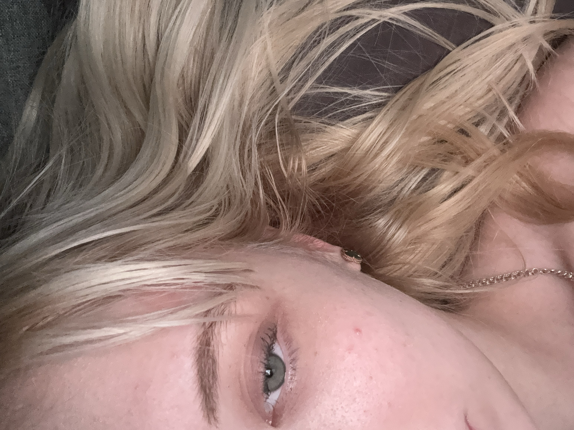A close-up, eye-level shot captures the left side of a person's face, focusing on their eye, cheek, and hair. The individual has light skin with a scattering of freckles and a single visible pimple on the cheek. Their eye is a striking green, with dark eyelashes and a faint hint of eyeshadow. A dark eyebrow arches above the eye. A simple gold chain necklace is visible around the neck, and a small earring with a dark stone is worn in the ear. The person's hair is a blonde, wavy texture, framing the face. The background is a dark, textured surface, possibly fabric. The lighting is soft, creating a gentle illumination on the skin.