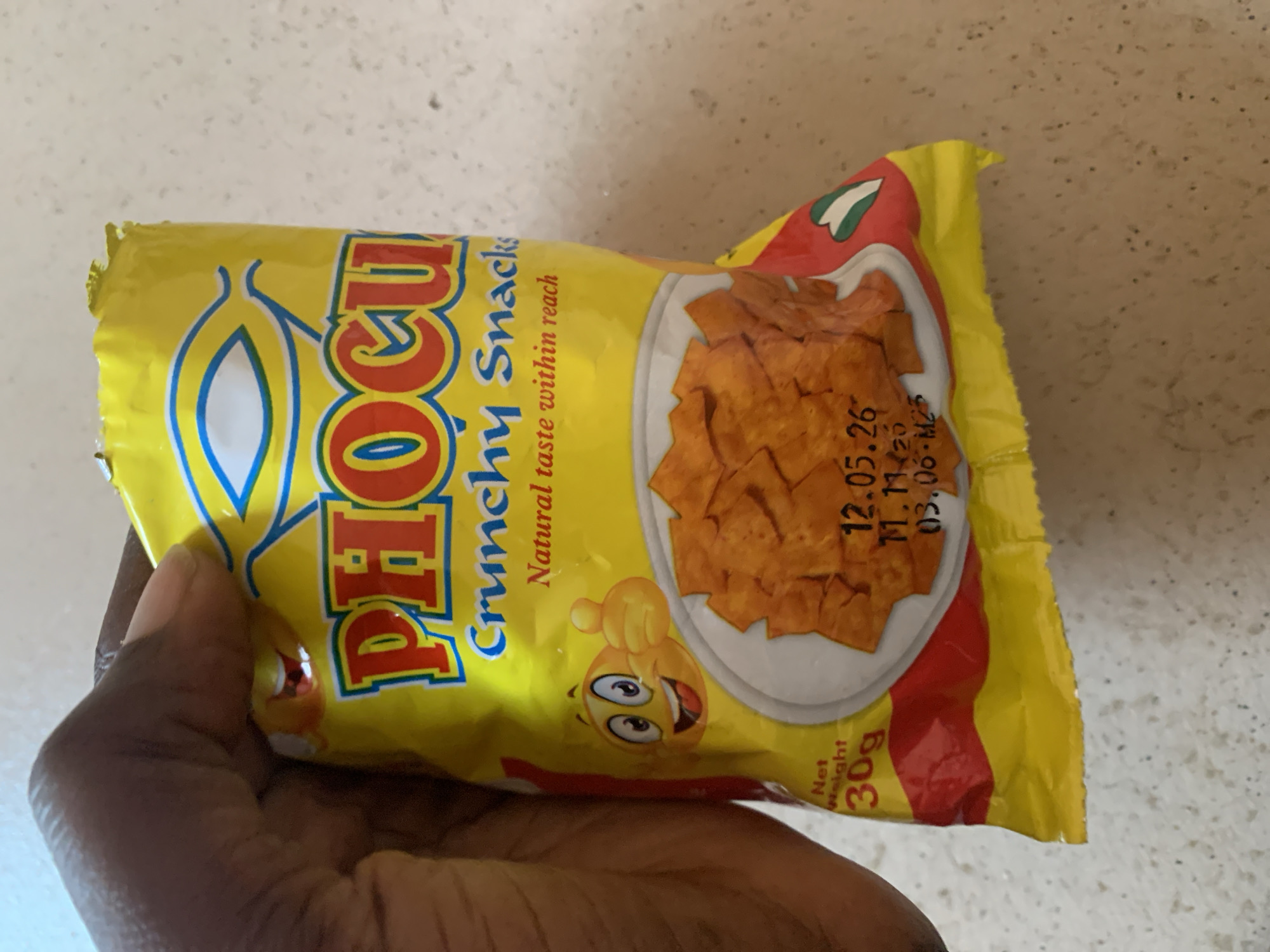 A hand holds a yellow snack bag with red and blue lettering that reads "PHOCU Crunchy Snacks". The bag features a cartoon emoji with wide eyes and a thumbs-up gesture, as well as an image of the snacks themselves. The text "Net Weight 30g" is visible on the lower left side of the bag, and a date "12.05.26" is printed on the right. The background is a speckled, light-colored surface.