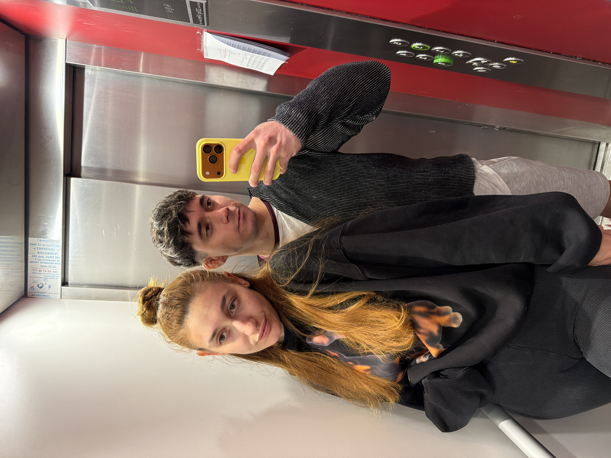 A man and a woman are taking a selfie in an elevator. The man is holding a yellow cell phone. The woman has red hair and is wearing a black sweatshirt with a design on the front. The man is wearing a dark long-sleeved shirt with light stripes. The elevator has stainless steel walls and a red panel with buttons. An advertisement with Cyrillic text is visible on the left side of the elevator wall. The location is likely in Sofia, Bulgaria, based on the Cyrillic text.