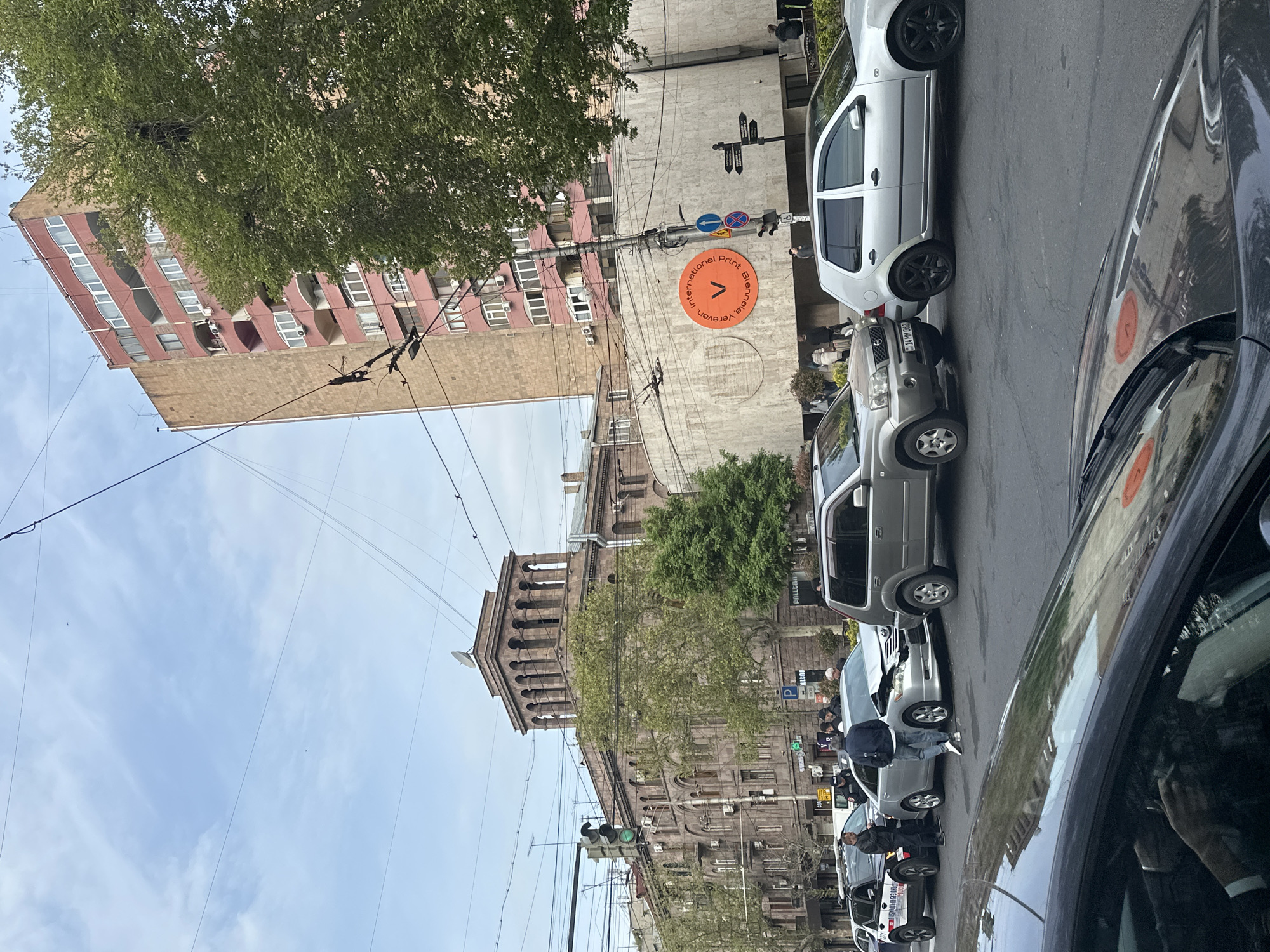 The image depicts a bustling urban street scene during the daytime, likely in Yerevan, Armenia, as indicated by a large circular sign. The sky is bright with some scattered clouds.
In the foreground and midground, multiple cars are parked along the side of the street. A dark vehicle occupies the immediate foreground, with its reflective surface showing reflections of the surroundings. A silver hatchback and a grey Nissan X-Trail (license plate "14 YN 060") are clearly visible. Further down the street, a silver car with noticeable front-end damage is present, around which several people are gathered on the sidewalk, suggesting a minor car incident or accident.
The street is bordered by various buildings. On the left, a tall, reddish-brown apartment building with numerous windows and balconies is partially obscured by lush green trees. Adjacent to it, a large, ornate stone building featuring arched windows and a prominent tower structure stands out. A modern grey building facade is also visible, hosting a distinctive orange circular sign that reads "International Print Biennale, Yerevan". Power lines and streetlights crisscross the scene.
Visible text includes "International Print Biennale, Yerevan" on the orange sign, "14 YN 060" on a car license plate, "P" on a parking sign, and "BOLERO" (along with its Armenian script "ԲՈԼԼԵՌՈ") on a building sign. Other partially visible text includes "ALLEON". Traffic lights are green, and other traffic signs indicate a one-way street and a no-stopping zone. The green foliage on the trees suggests the season is spring or summer.
