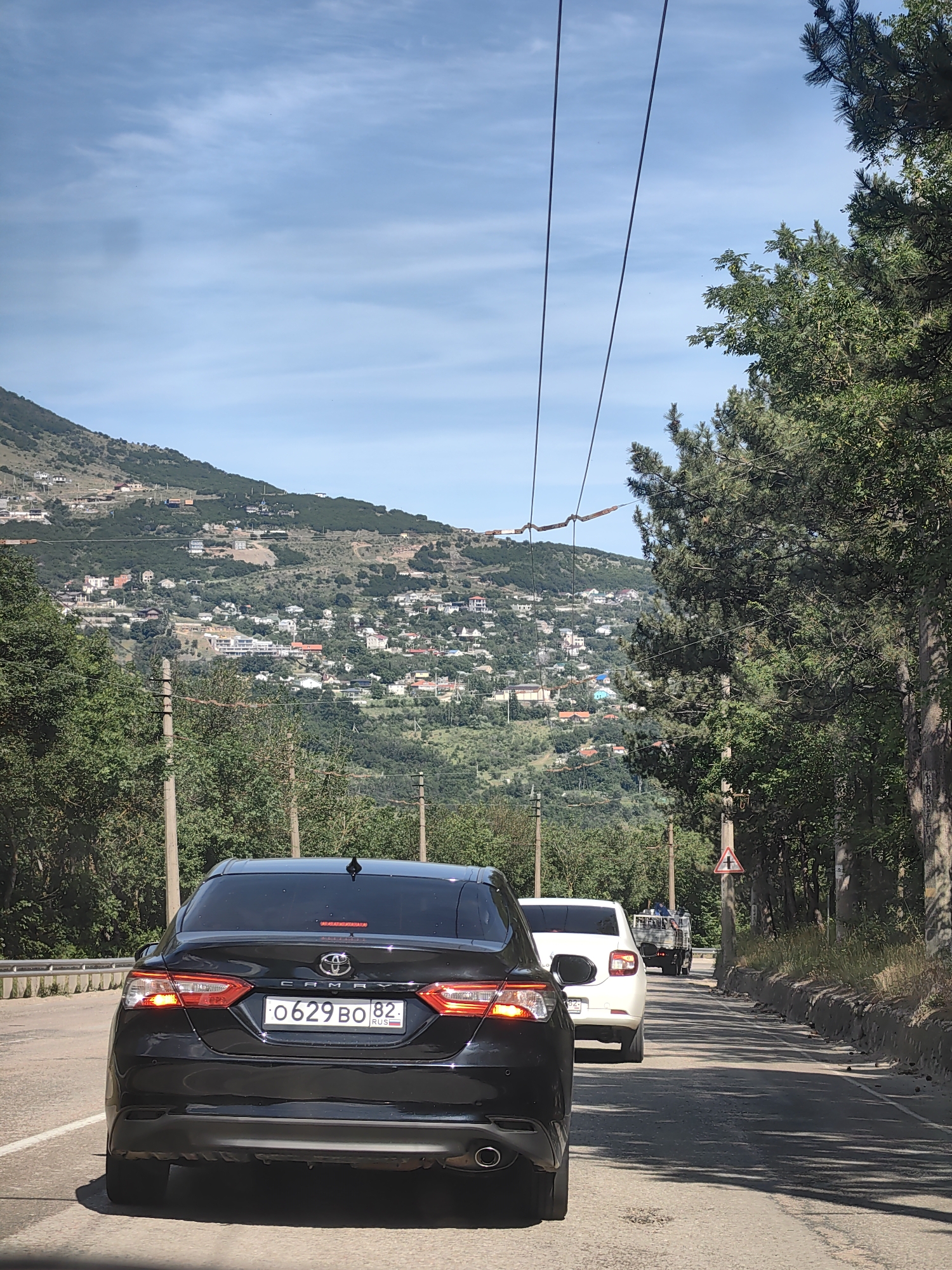 The image shows a view from a vehicle traveling on a road with a mountainous landscape on the left and a dense forest on the right. The road is paved and appears to be a two-lane road with a guardrail on the left side. Several cars are visible on the road, including a black Toyota Camry in the foreground, a white car behind it, and a utility vehicle further ahead. The mountainous area is dotted with numerous houses and buildings, suggesting a settlement. Power lines are strung across the sky, supported by utility poles. The weather appears to be clear and sunny, with a blue sky and some scattered clouds. The scene is consistent with a rural or suburban area near a coast or mountains, likely in the region of Kiparisnoe, Ukraine, given the context.