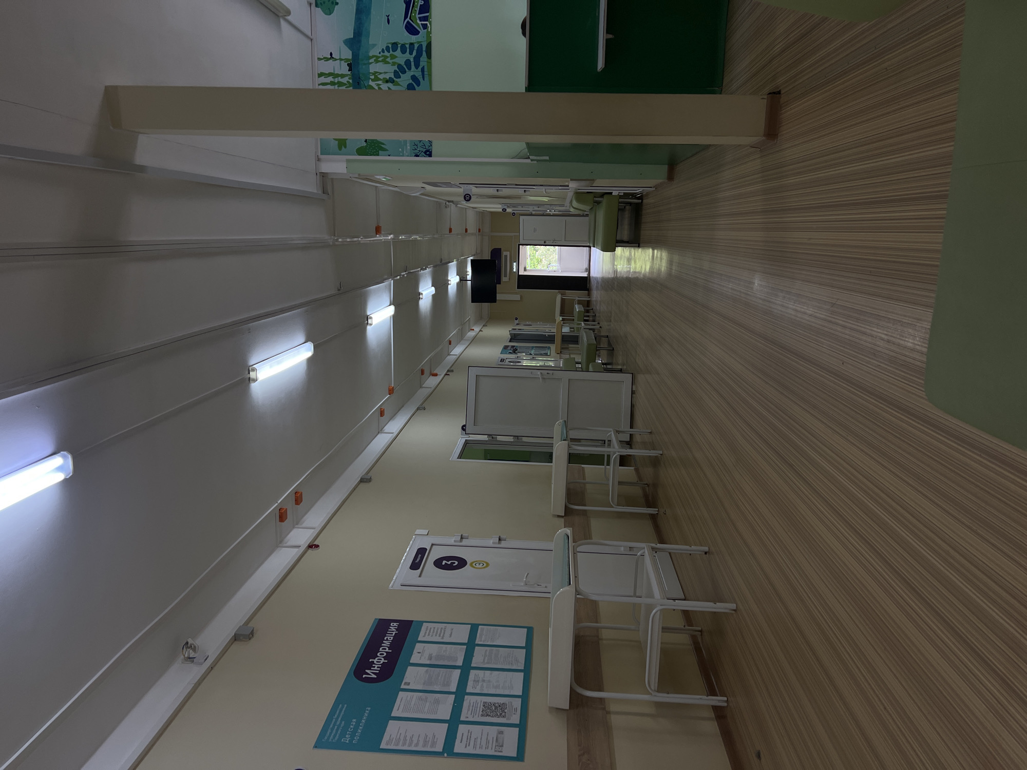 The image depicts a brightly lit, long hospital corridor. The walls are painted a pale yellow, and the floor is covered with light brown wood-patterned linoleum. Fluorescent lights are installed on the ceiling, illuminating the space. On the left side of the corridor, an information board is mounted on the wall, featuring various documents and what appears to be a QR code. Below the information board are two white metal tables with cushioned surfaces, resembling changing tables or medical examination beds. Further down the corridor, several doors are visible, some with room numbers displayed. One door has a prominent yellow circle with the number "3" inside. Green benches are placed along the right wall of the corridor. In the distance, natural light streams in from an open doorway, suggesting an exit or an outdoor area. The environment appears clean and sterile, typical of a medical facility. The image does not provide specific visual cues to confirm the location as Kuyeda, Russia, but the text on the information board appears to be in Russian, hinting at its location.