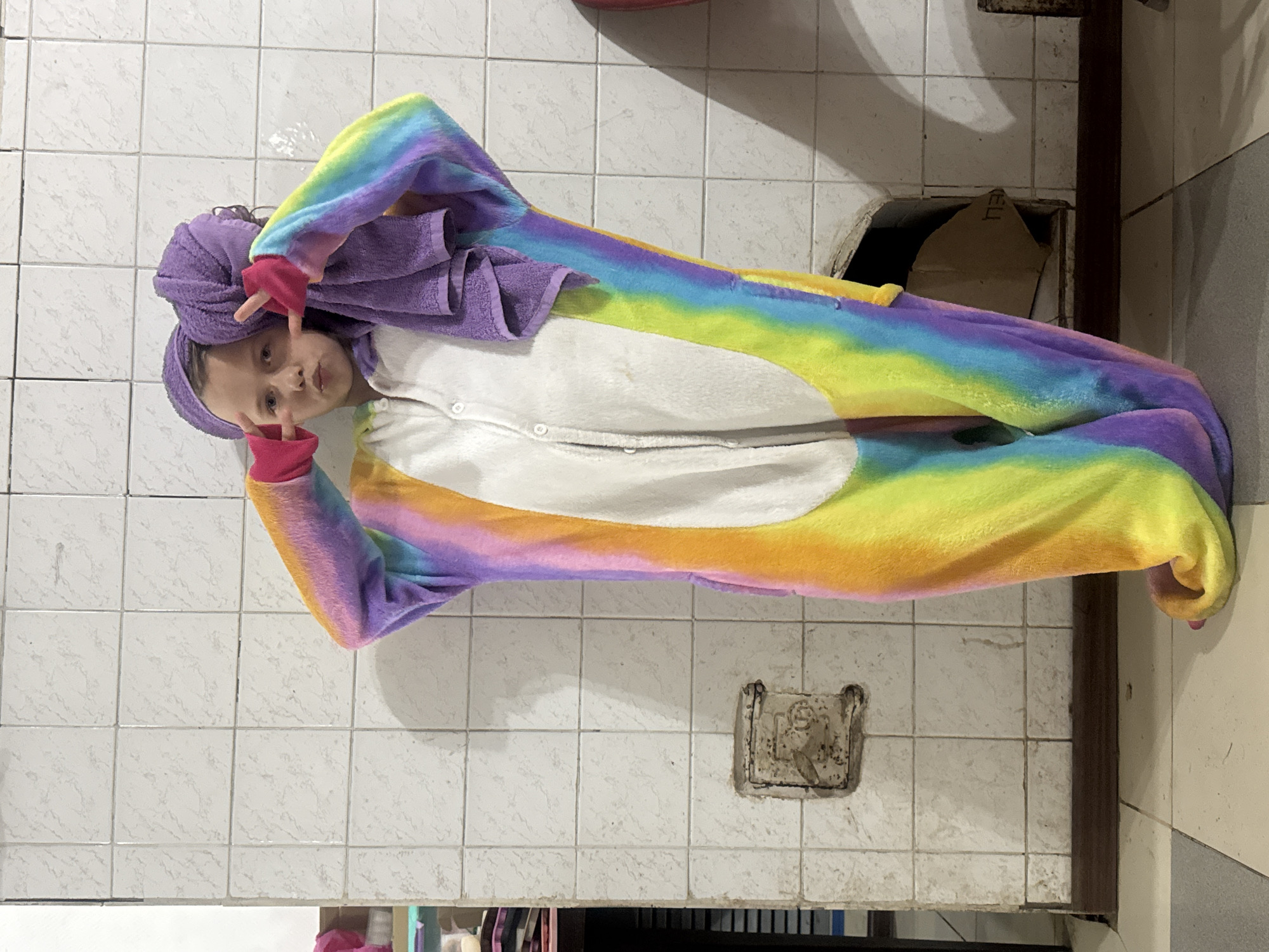 A young girl with a purple towel wrapped around her head is wearing a colorful rainbow unicorn onesie in a bathroom setting. She is making a peace sign with both hands. The bathroom has white tile walls and what appears to be a utility access panel in the wall. There is also a remnant of a wall-mounted accessory, possibly a soap dish or a toothbrush holder. The precise location within the city of Rusalevka, Russia, cannot be determined from the image.