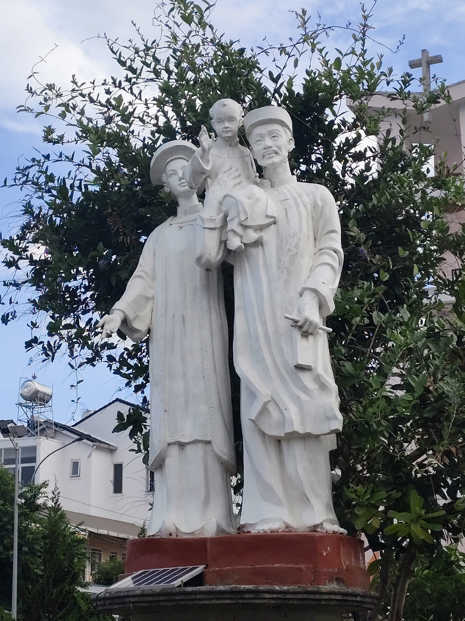 The image depicts a white marble statue of the Holy Family, which is a sculpture of Saint Joseph holding the Child Jesus on his shoulders while his wife, the Virgin Mary, stands next to him. The statues are dressed in Vietnamese traditional clothing. The statue stands in front of greenery, trees, and plants on top of a red base, with a solar panel affixed at the bottom. In the background, there is a white building, presumably a church, with a cross visible on the roof. Other buildings and a water tower are also visible in the background. The image appears to be taken in Da Lat, Vietnam, based on the style of the statue and the surrounding environment.