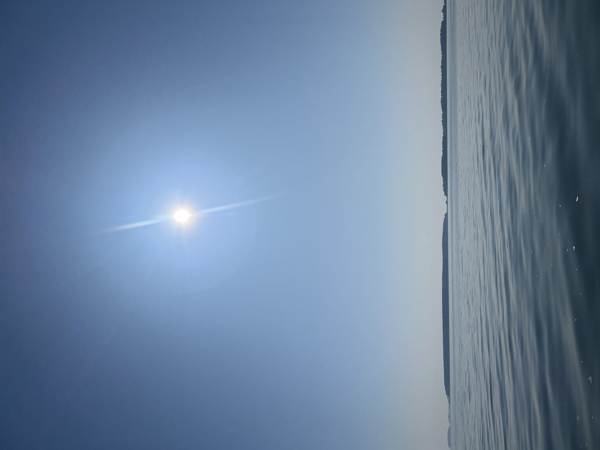 The image captures a serene body of water under a clear blue sky, with the sun shining brightly and emitting a lens flare. In the distance, a long, dark, tree-lined island stretches across the horizon. The water's surface is gently rippled, reflecting the light and creating a textured pattern. There are no people, animals, or structures visible in this scene. The overall impression is one of peacefulness and natural beauty, evoking a sense of openness and tranquility. The scene is likely set in a natural environment, and it is not possible to determine a specific location within the city of Novopichugovo, Russia, based on the visual information provided.