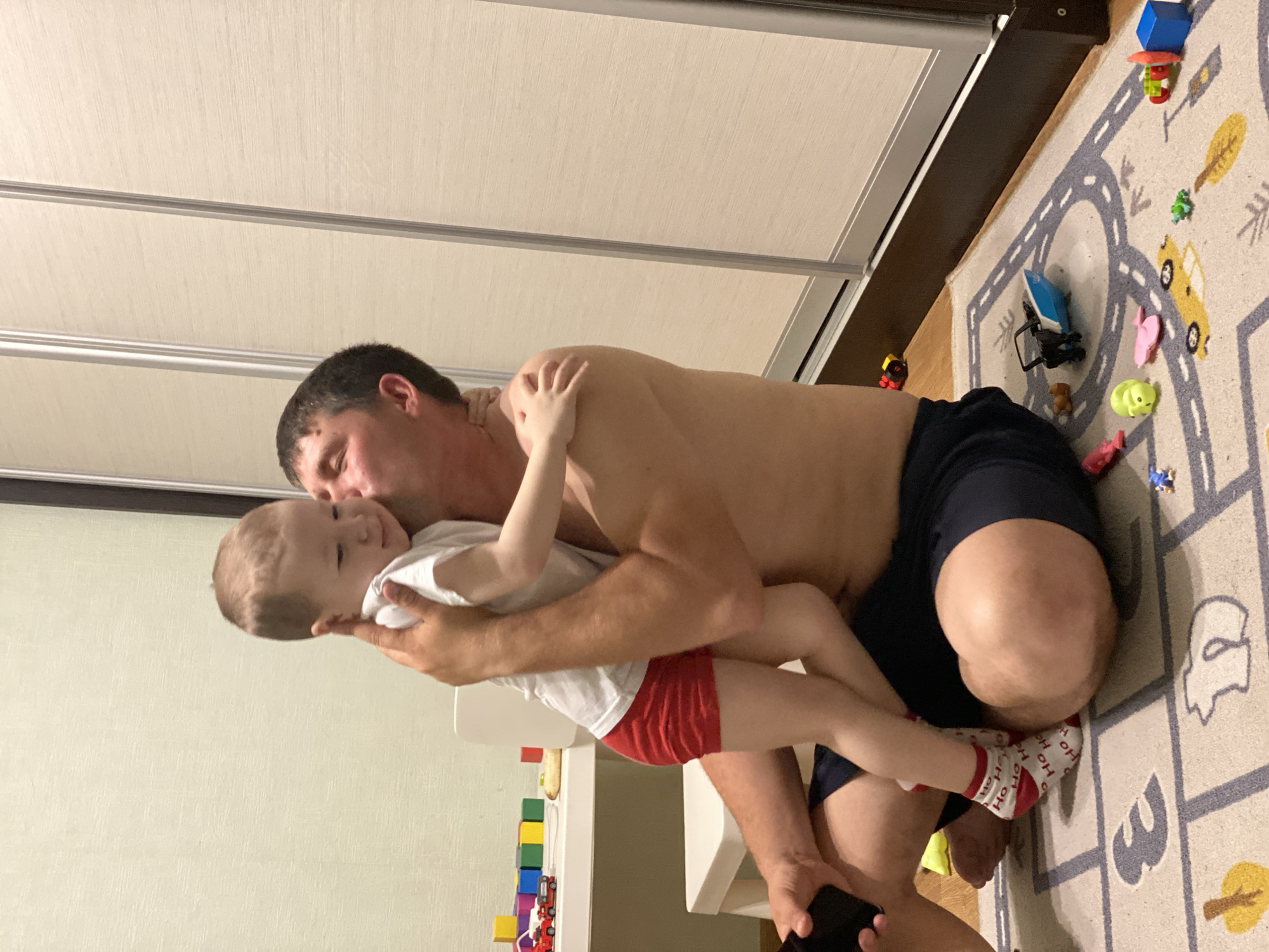 This image, taken in Sochi, Russia, captures an affectionate indoor moment between an adult male and a young child. The adult, presumably the father, is shirtless and wearing dark shorts, kneeling on a patterned rug. He is seen tenderly kissing the young child on the cheek. The child, likely a boy, is held in the father's arms, wearing a white t-shirt, red shorts, and white socks with red stripes and the text "HO HO HO." His left arm is gently draped around the father's neck, and he has a calm, content expression.
The setting appears to be a child-friendly room or play area. A light-colored rug featuring a road and town design covers the floor, populated with numerous colorful toys including building blocks, toy cars, small animal figures, and a blue toy boat. A white piece of furniture is visible to the left of the child, with a stack of multi-colored building blocks on the floor nearby. In the background, there is a light-colored wall and a large closet or wardrobe with light-paneled sliding doors and metallic frames. The indoor lighting suggests the photo was taken in the evening or at night. The scene conveys a strong sense of love and warmth between the father and child.