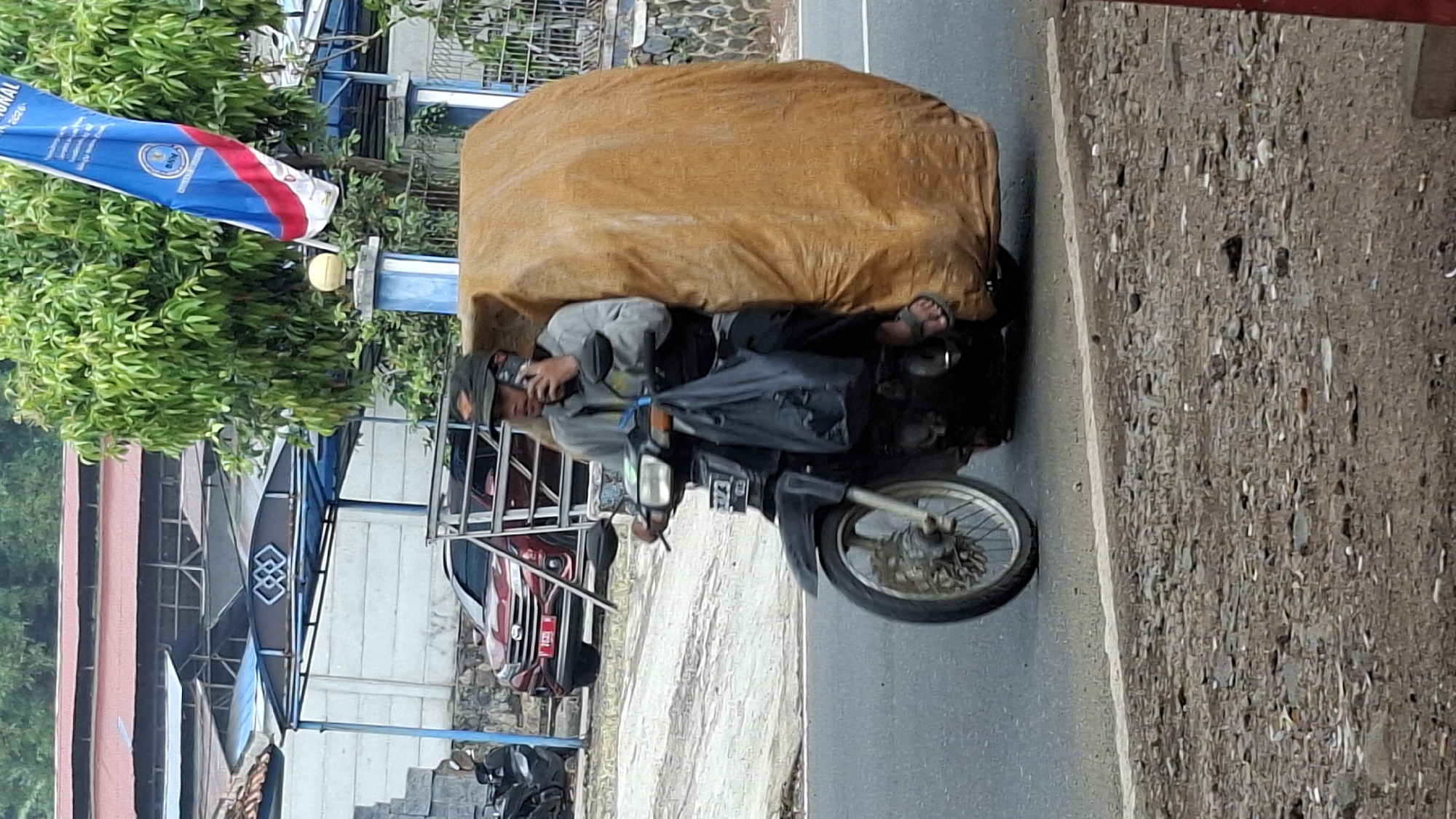 In Cilaku, Indonesia, a man rides a black motorcycle down a paved road, talking on his phone. He is wearing a grey long-sleeved shirt, black pants, and a dark cap. The motorcycle carries a large, bulky object wrapped in a tan tarp on its rear. A black plastic bag hangs from the handlebars. In the background, buildings with red and blue roofs and a blue and white banner are visible. A red car is parked on the left side of the road. The environment appears to be a semi-urban or rural area with trees and some rough terrain alongside the road.