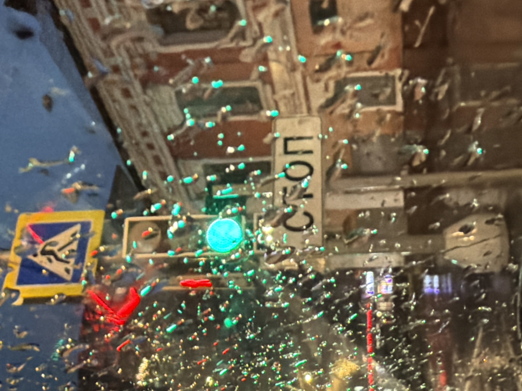 A traffic light with a green signal is visible. A street sign with the word "СТОП" (STOP) in Cyrillic script is also present. A pedestrian crossing sign is in the background. The scene is illuminated by streetlights and possibly car headlights. The image appears to have been taken through a wet window, with numerous water droplets obscuring parts of the view. The overall impression is that of a city street at night, possibly during or after rainfall. No specific landmarks that definitively place it in Moscow, Russia, are identifiable from the provided image.