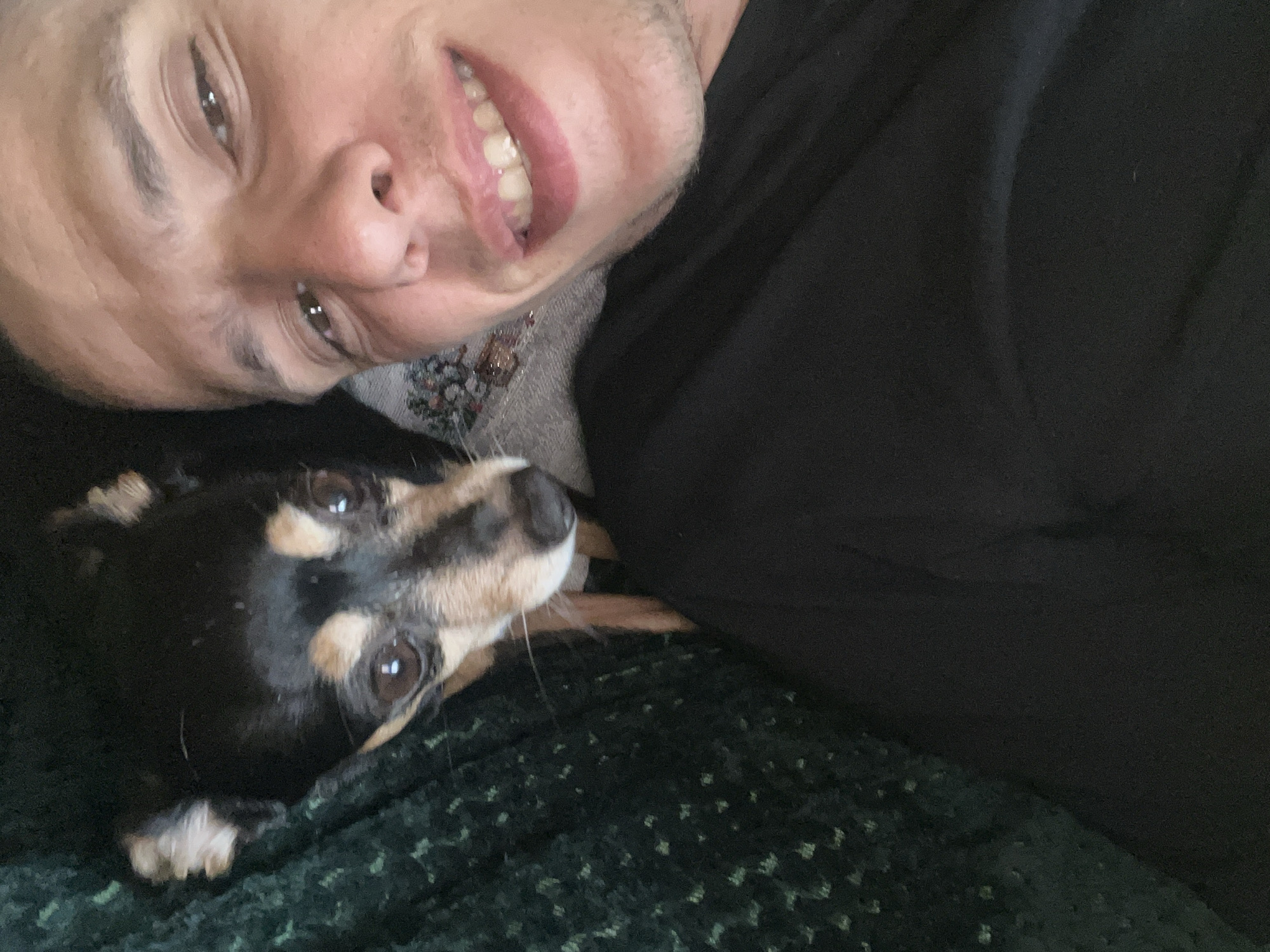 A person and a small dog are in the image. The person, likely male, is smiling at the camera. He has short dark hair, tan skin, and is wearing a dark top over a gray shirt with a floral design near the neck. The dog is black and tan with short hair and is lying near the person. They are on a dark green textured surface, possibly a couch or blanket. The image does not contain enough information to establish that it was taken in Sofia, Bulgaria.